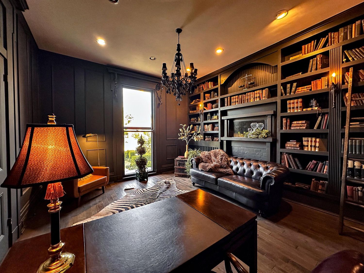 Dark luxury home library with leather Chesterfield sofa, floor-to-ceiling bookshelves, chandelier, and cowhide rug.