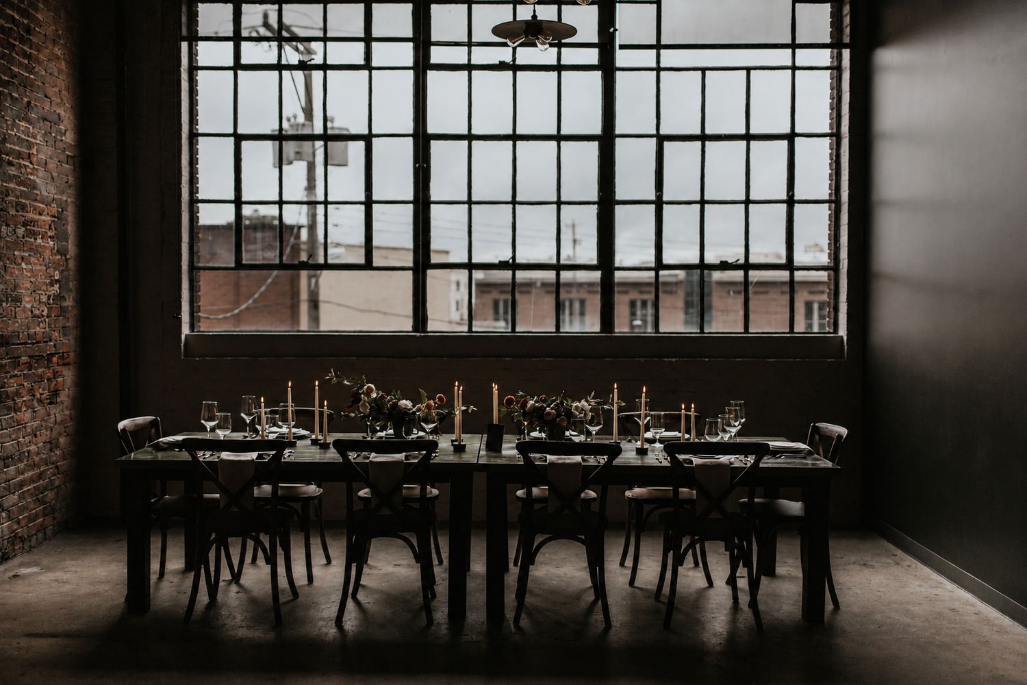 Elegant industrial dining setup with long black table, candles, and chairs framed by large warehouse windows.