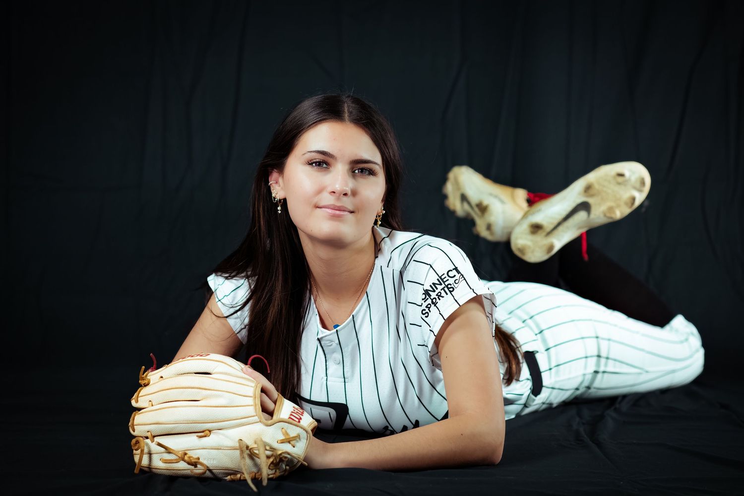 Young female softball player lying down smiling with glove in pinstripe uniform.