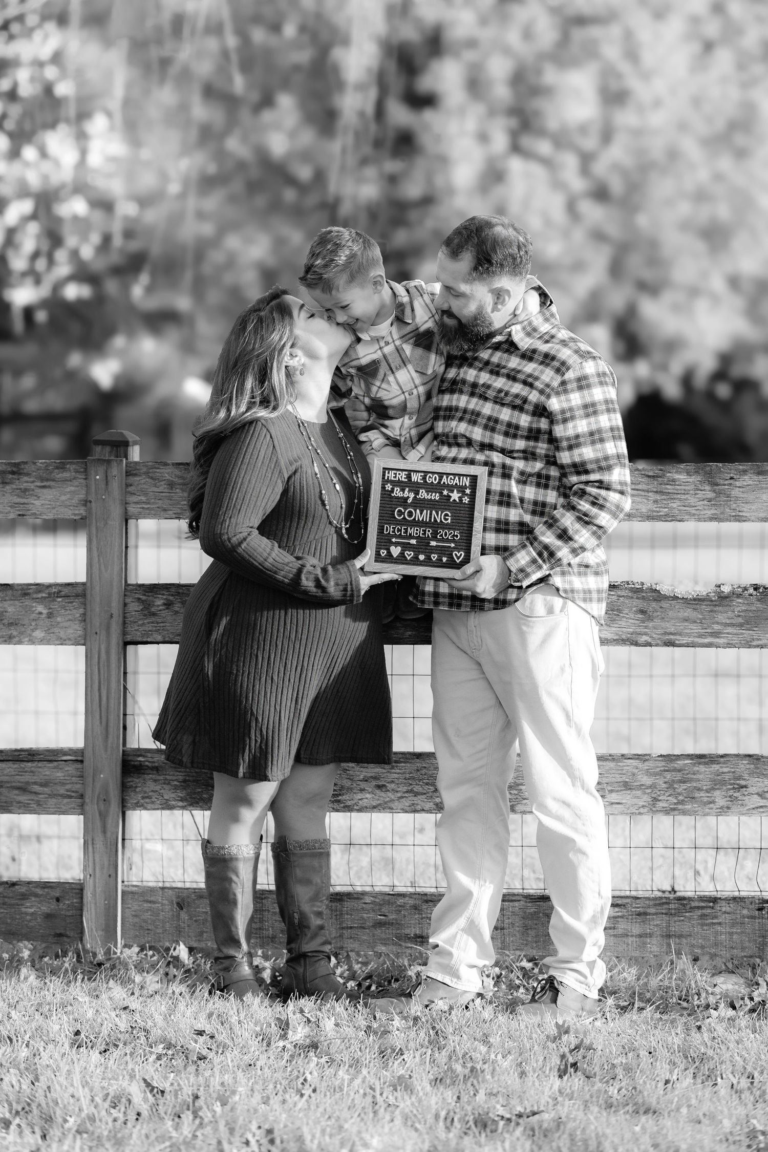 Couple and friend holding pregnancy announcement sign by rustic fence in black and white maternity photo.