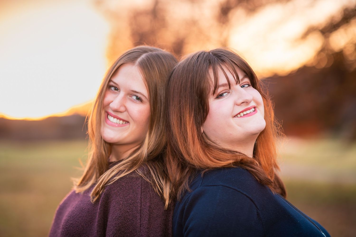 Two young women smiling back to back during a golden hour outdoor portrait session.
