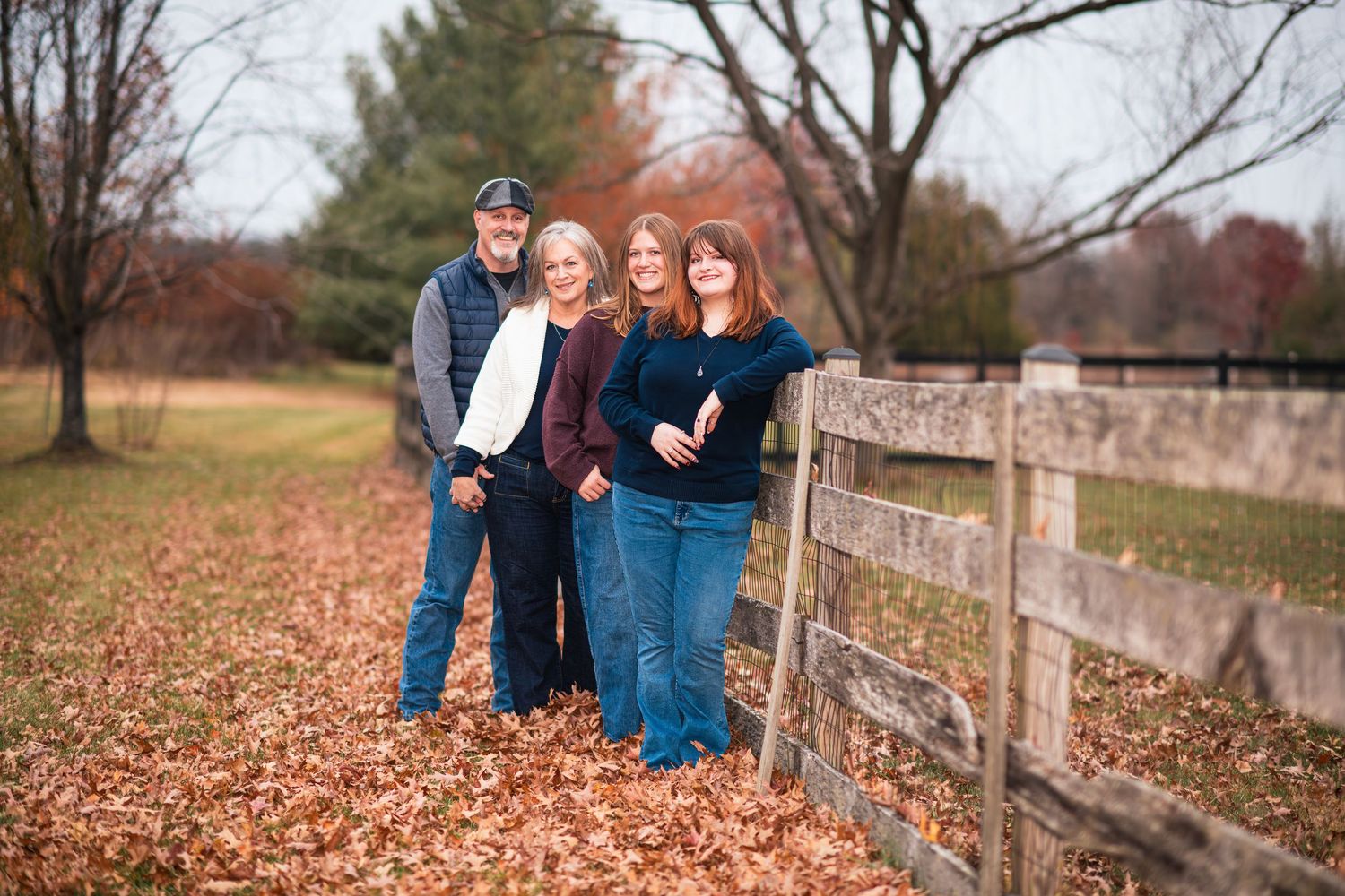 Family of four posing along rustic wooden fence in colorful autumn leaves.