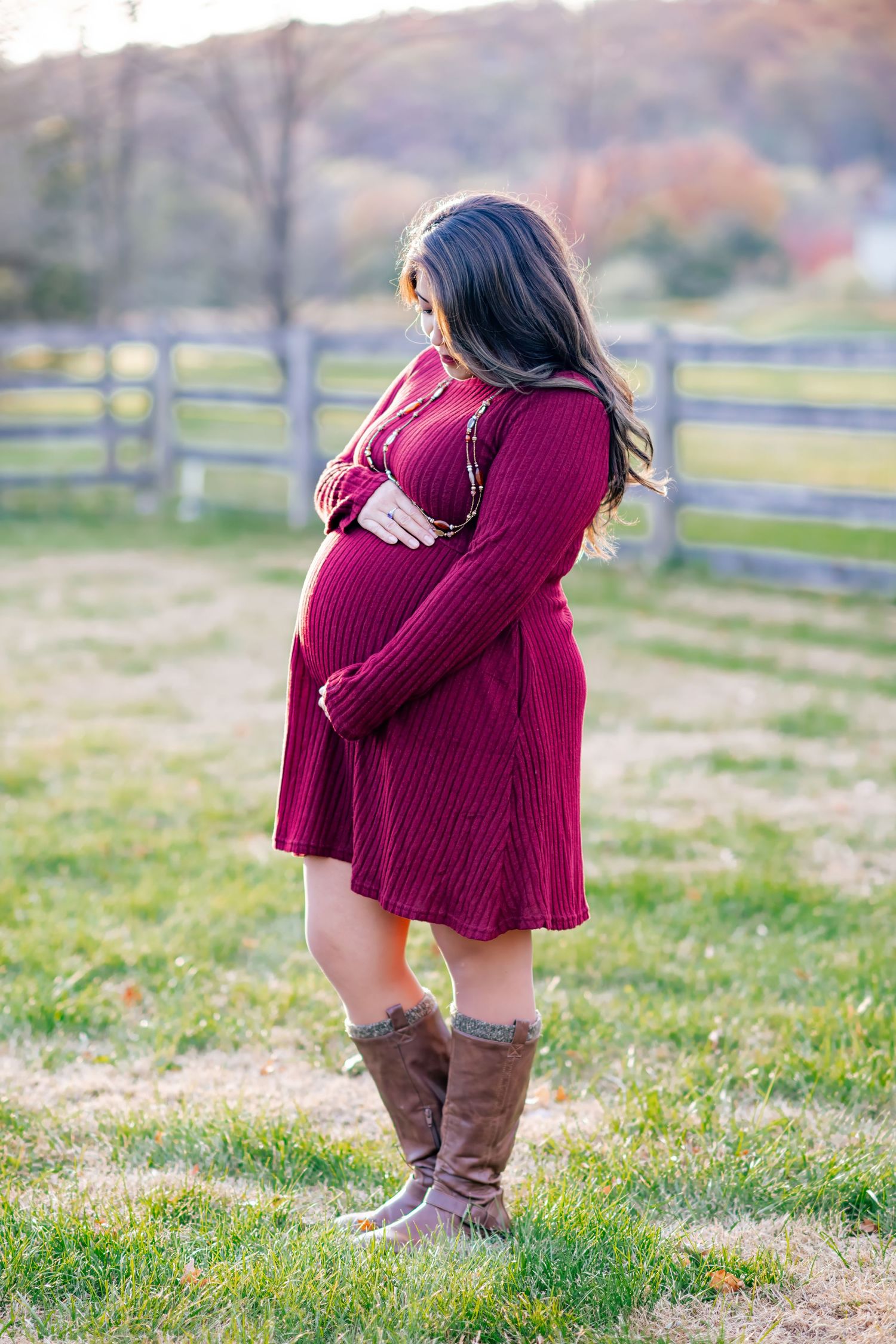 Pregnant woman in burgundy dress and boots cradling baby bump in autumn field.