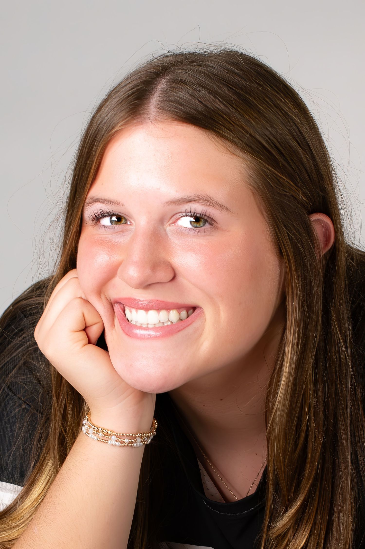 Smiling teenage girl with long brown hair resting chin on hand wearing bracelet.