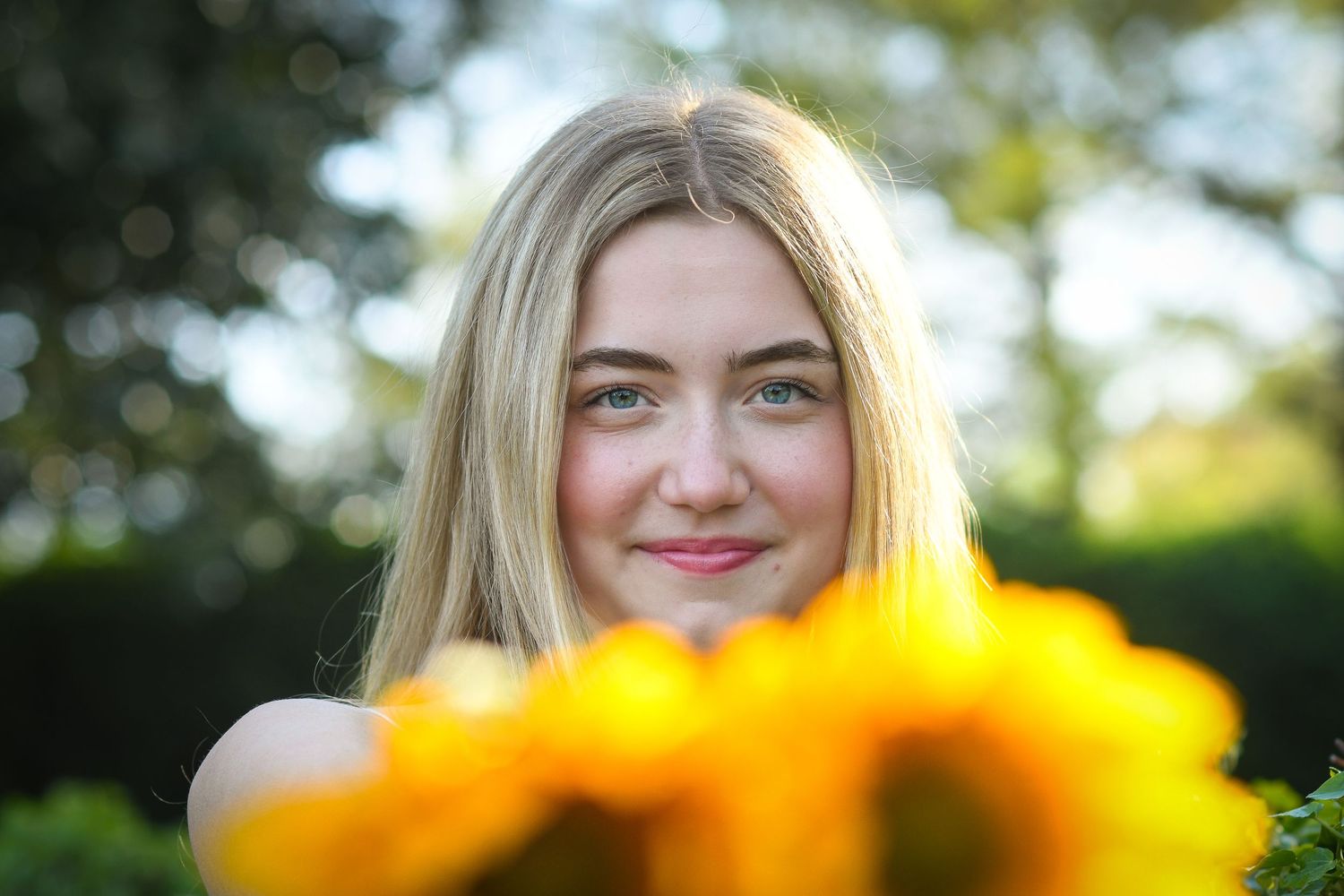 Smiling blonde girl peering over bright yellow sunflowers in a sunny garden portrait.