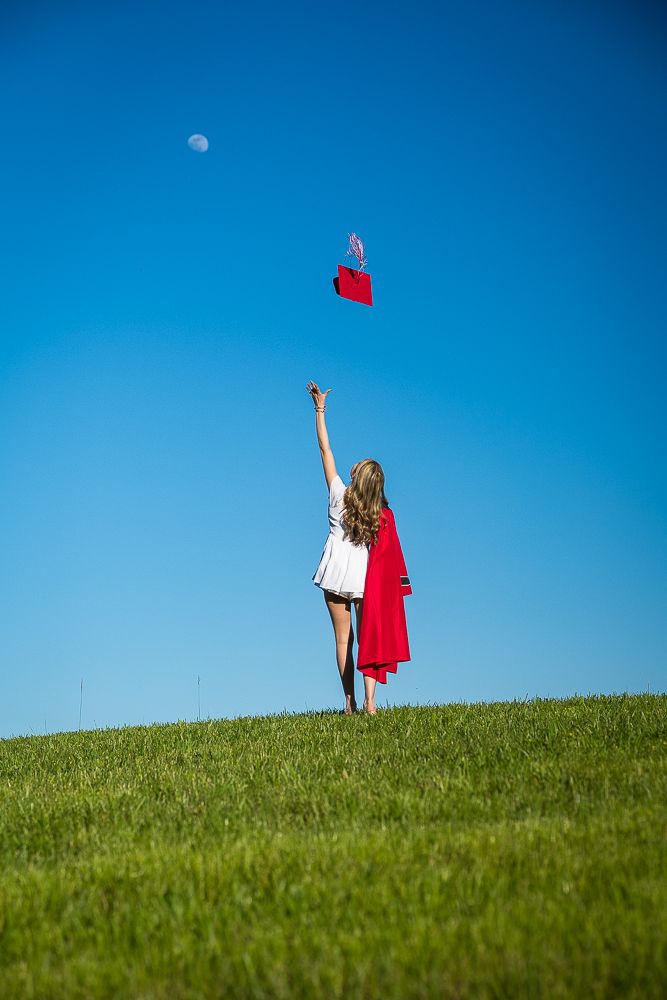 Girl in white dress tossing red graduation cap into blue sky on green hill with moon visible.