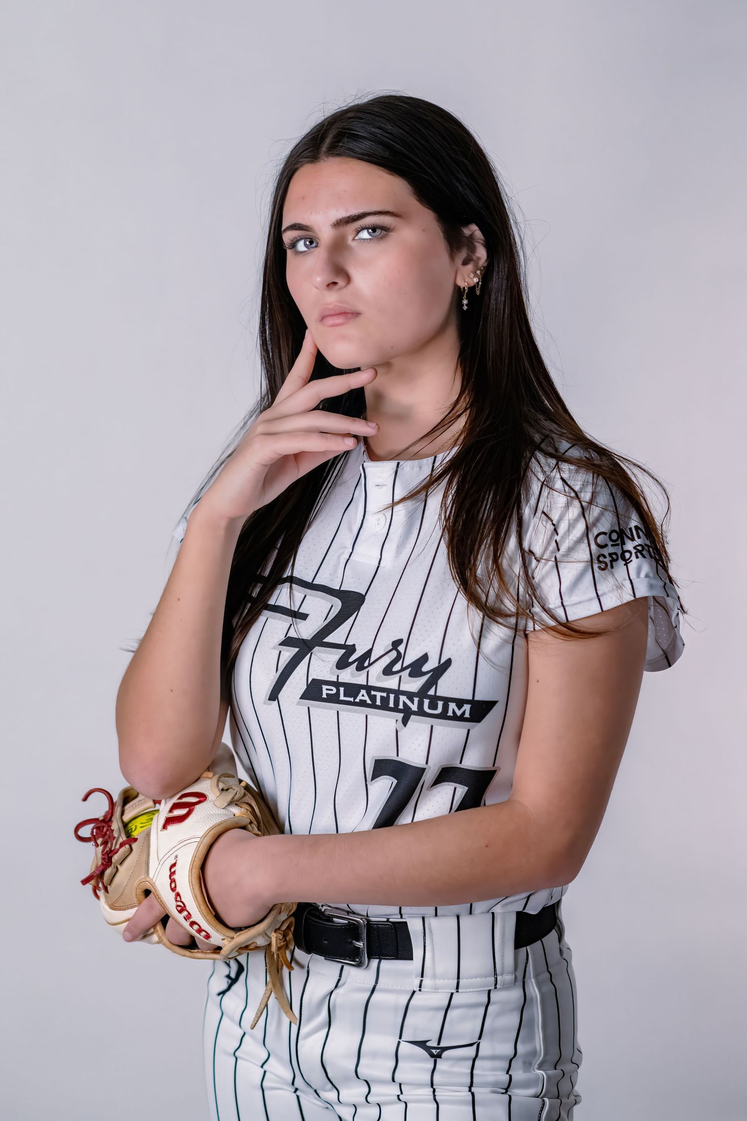 Female softball player #77 in Fury Platinum pinstripe uniform posing thoughtfully with glove.