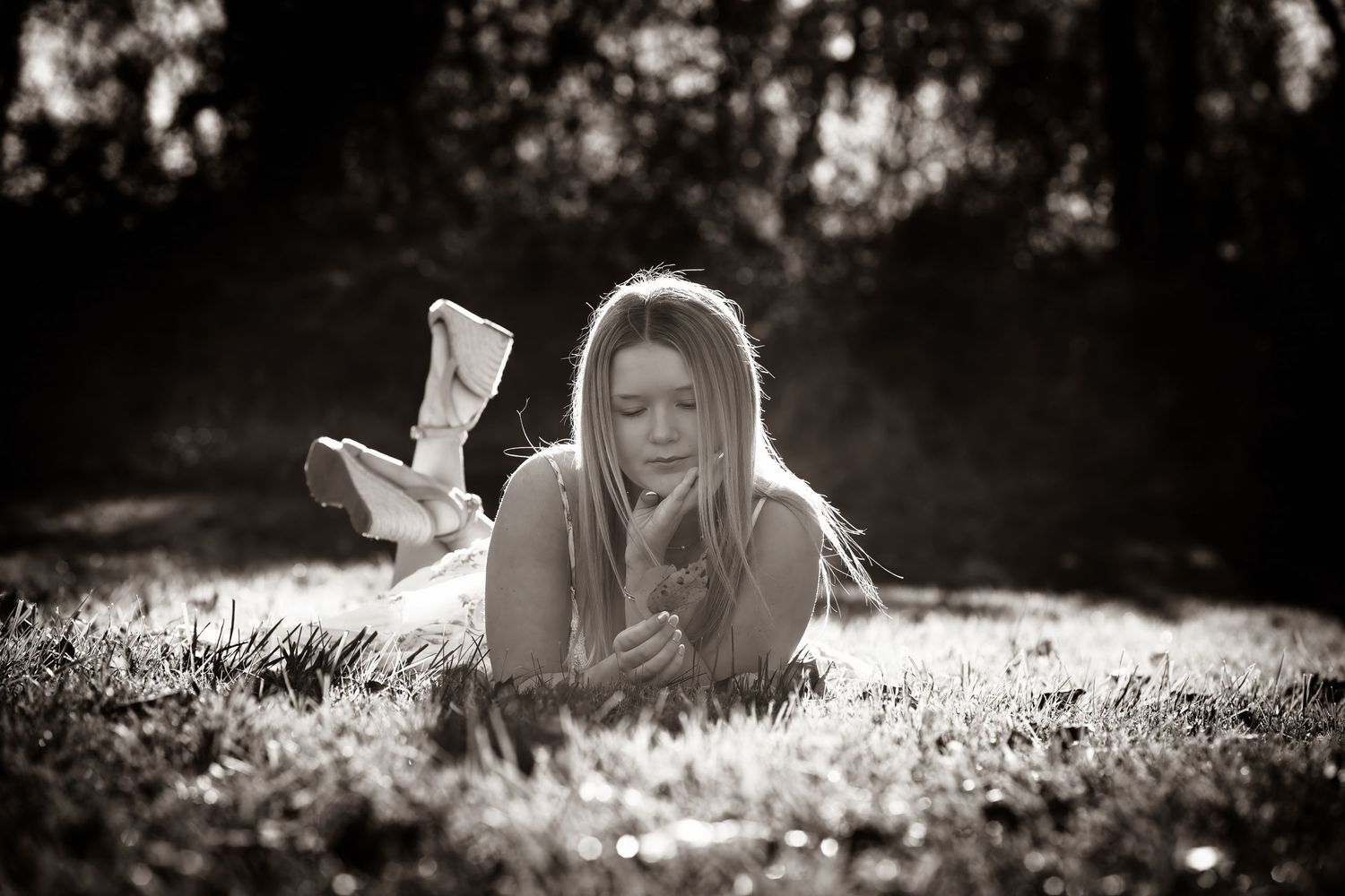 Young blonde woman lying on grass in sunlight, holding a flower, dreamy black and white outdoor portrait.