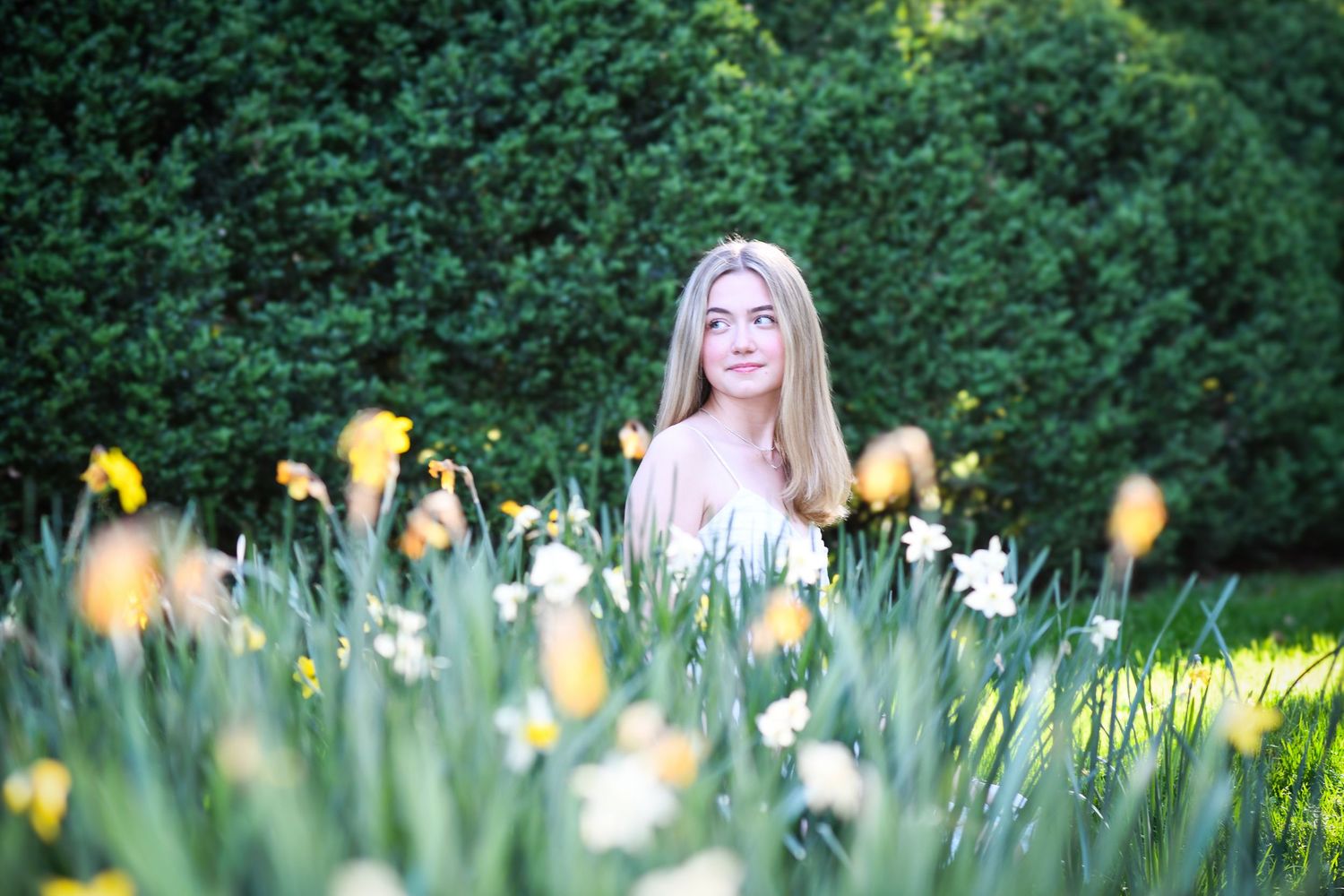 Young blonde woman sitting among white and yellow daffodils in a lush green garden.