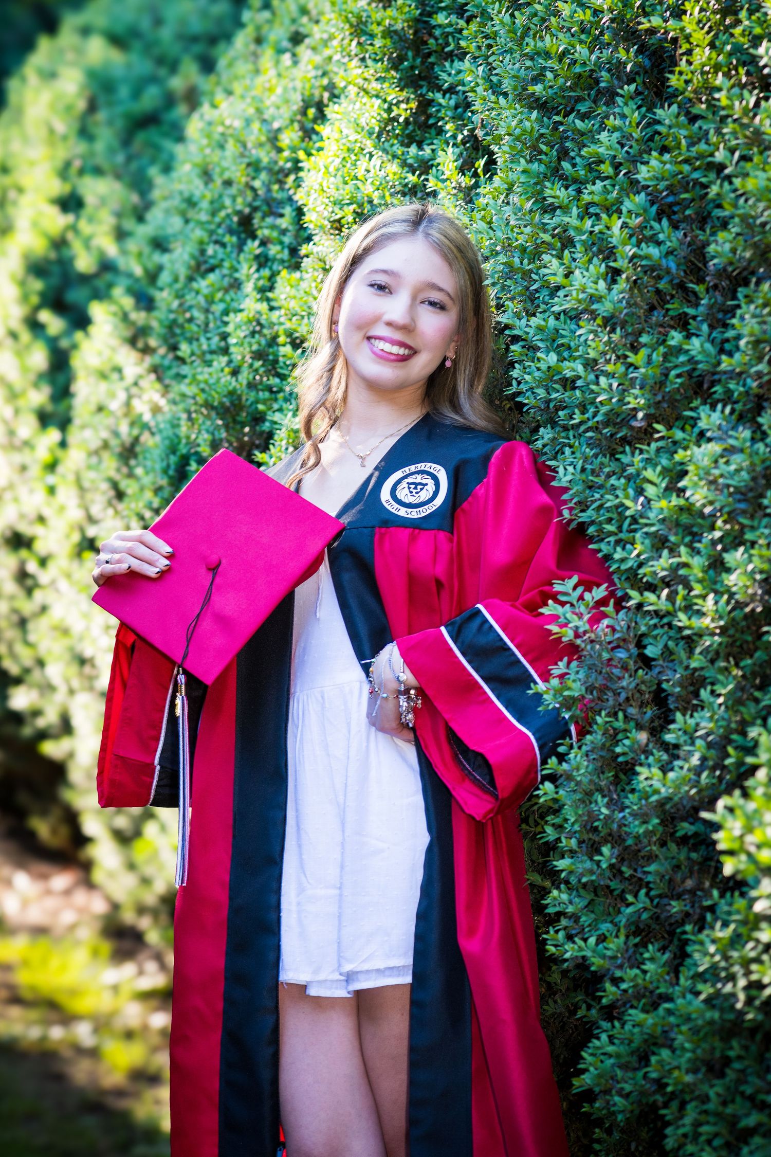 Smiling graduate in red and black graduation gown holding cap among green hedges for senior photos.