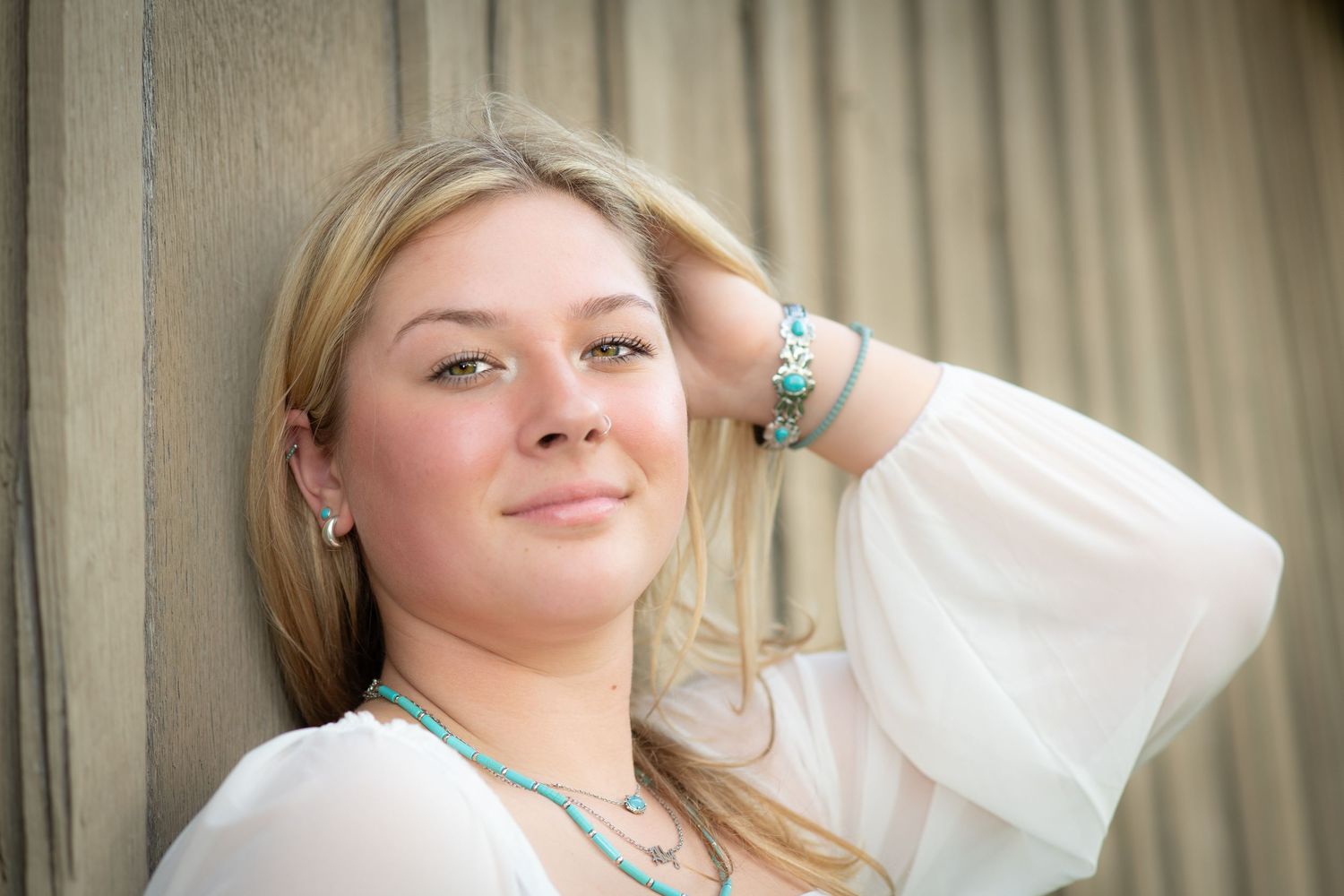Blonde woman in white blouse with turquoise jewelry poses against wooden wall.