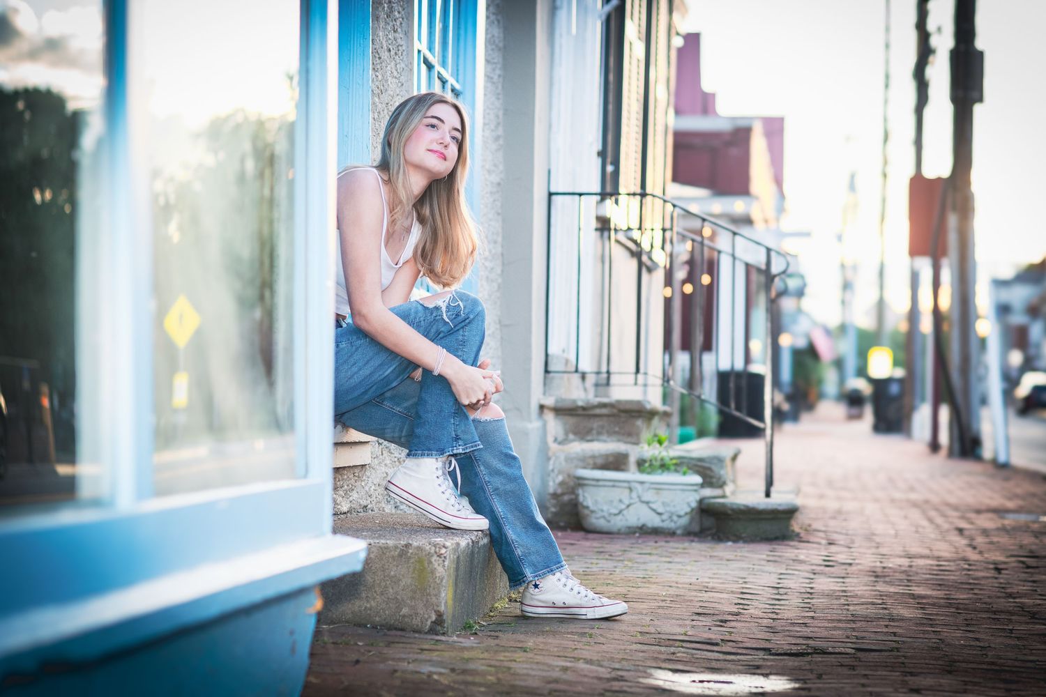 Young woman in jeans and tank top sits on storefront steps enjoying sunny urban street scene.