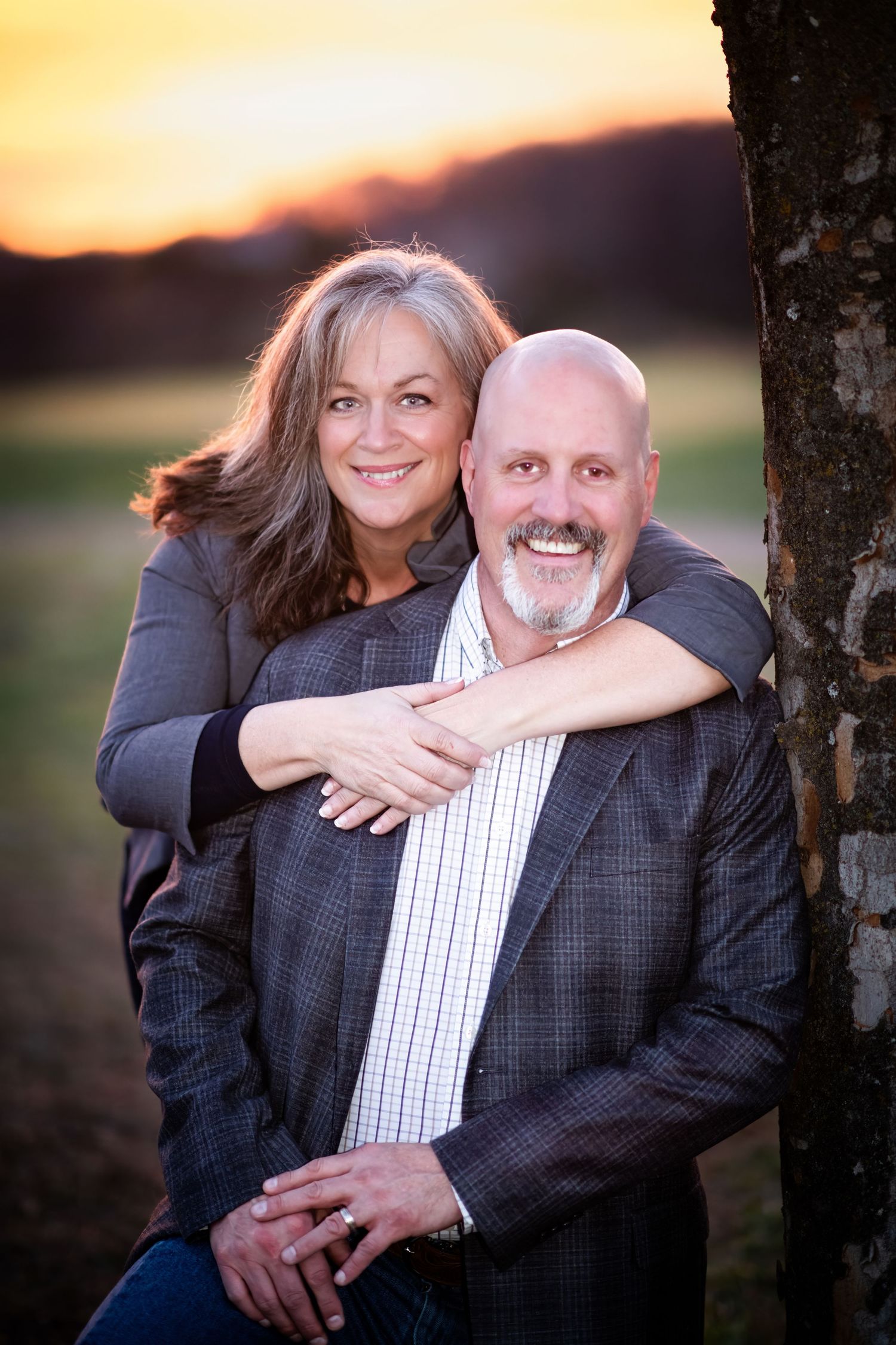 Smiling couple posing together at sunset, woman embracing man from behind near a tree.