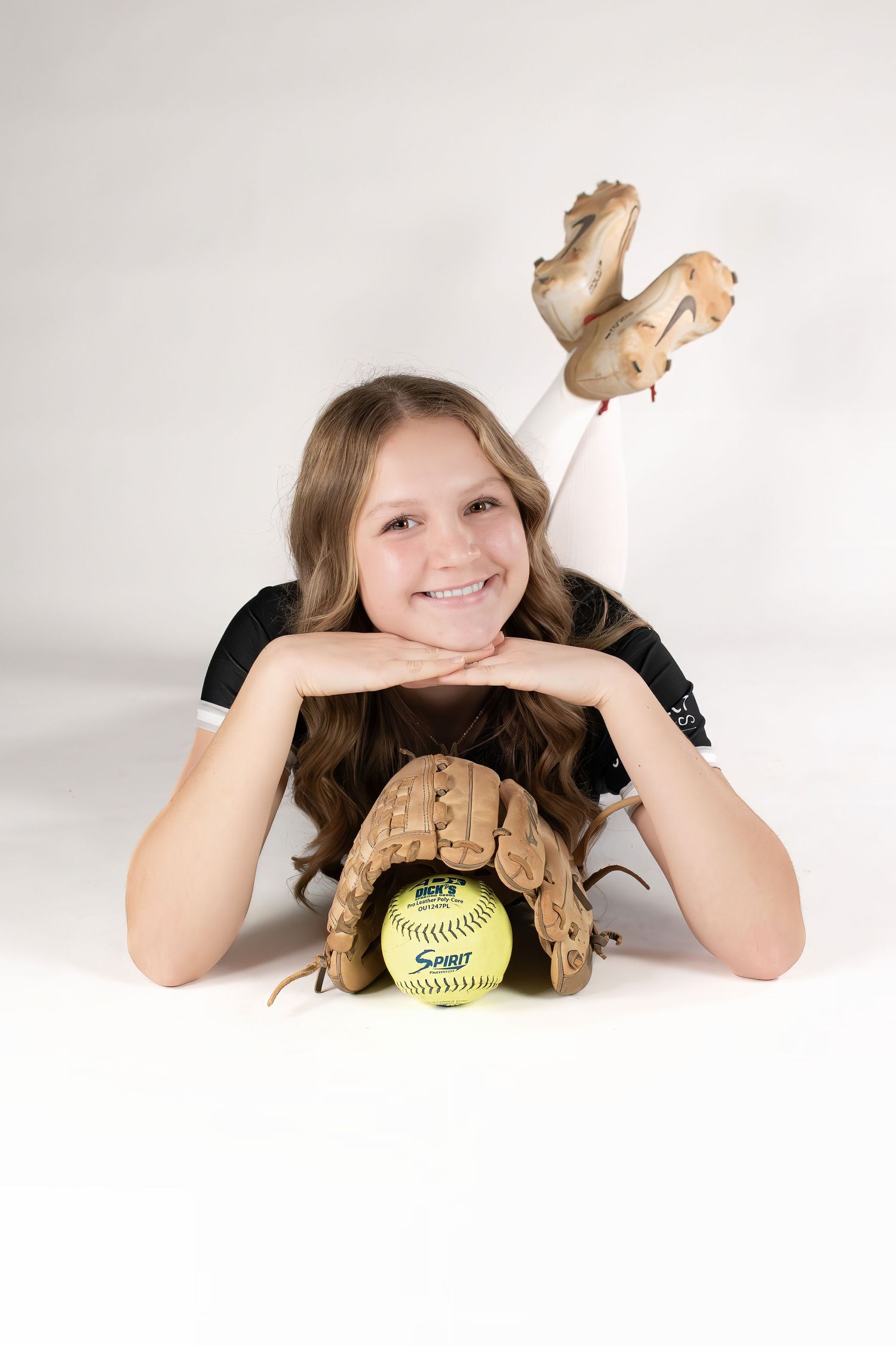 Smiling girl in softball uniform lying down with glove and yellow softball.