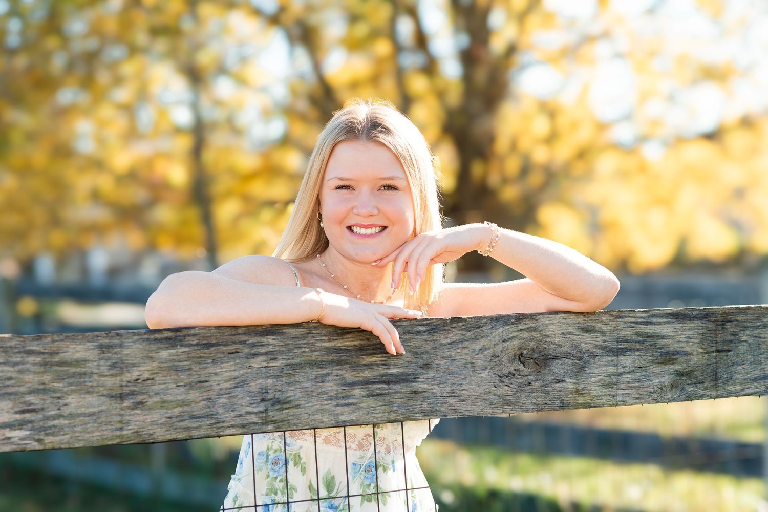 Smiling blonde girl leaning on rustic wooden fence in autumn sunlight.