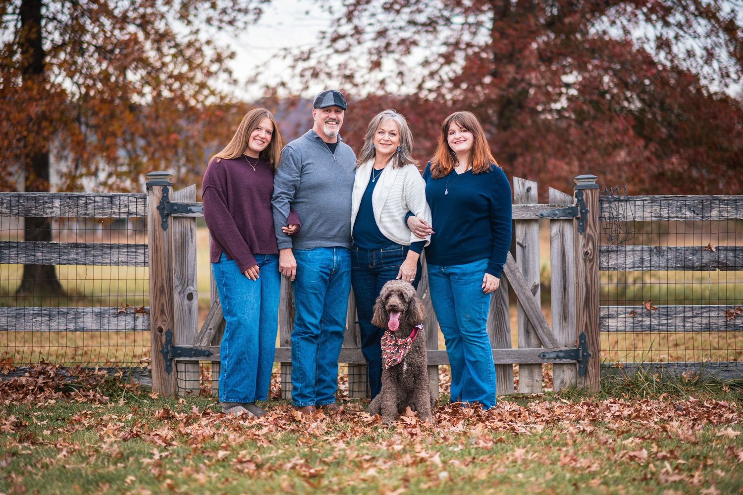 Family of four with dog posing by wooden gate surrounded by fall foliage.