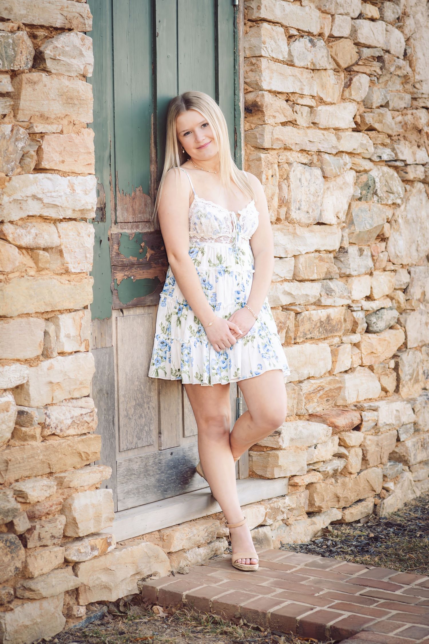 Blonde girl in floral sundress leaning against rustic stone wall by vintage door for senior photos.