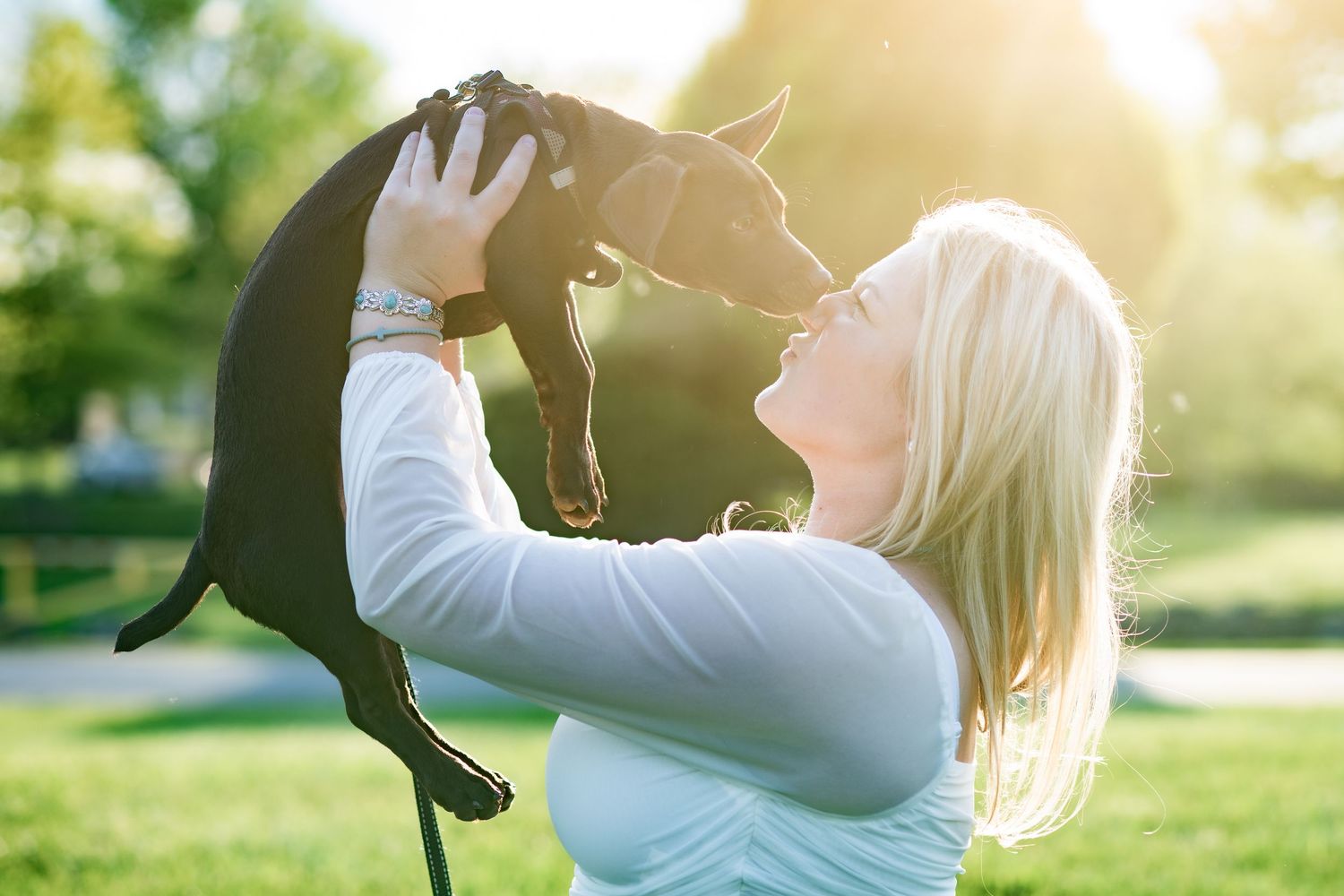 Blonde woman joyfully lifting a small black dog in a sunny green park, sharing a playful moment outdoors.