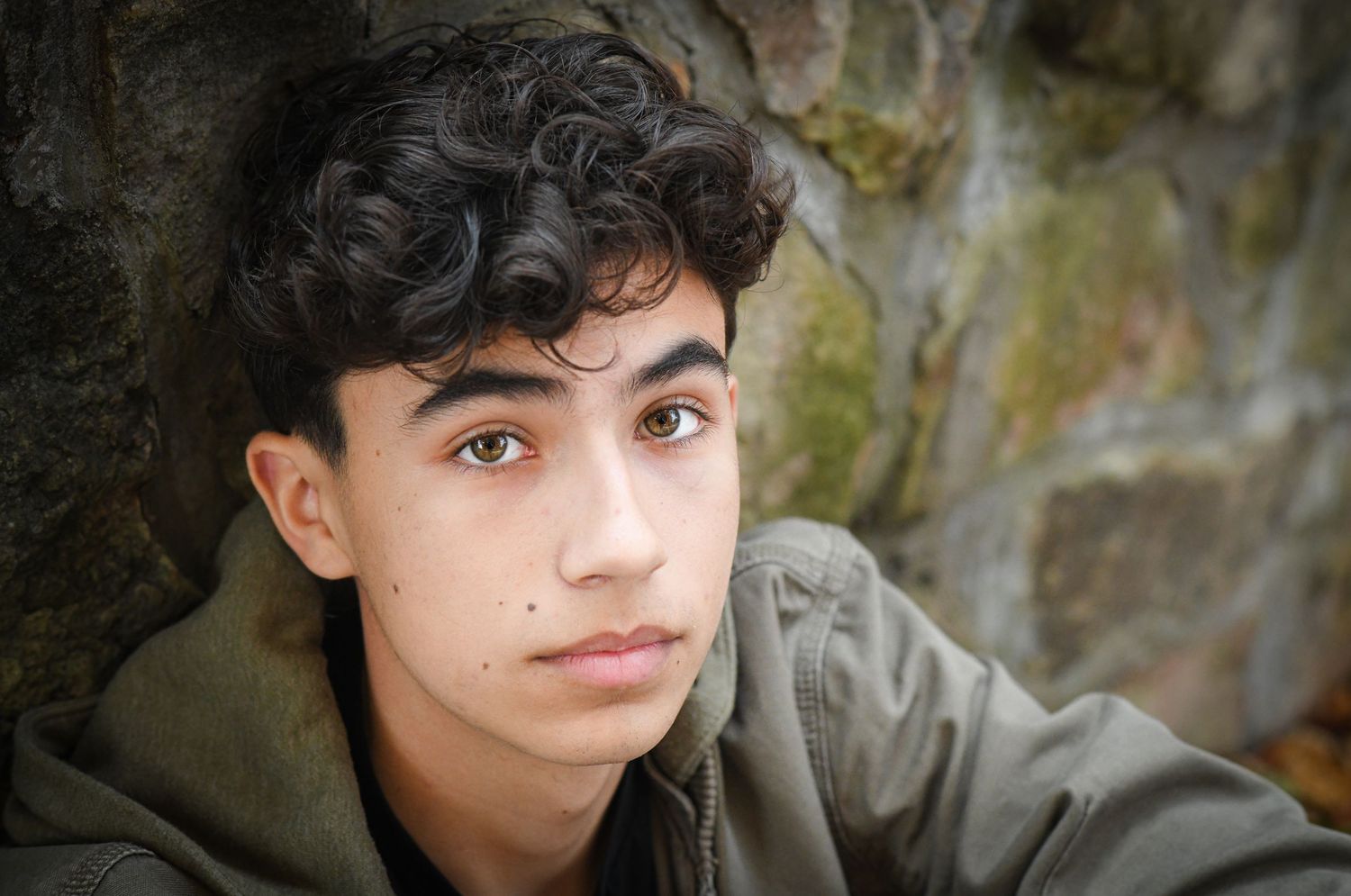 Teen boy with curly hair leaning against stone wall in jacket.