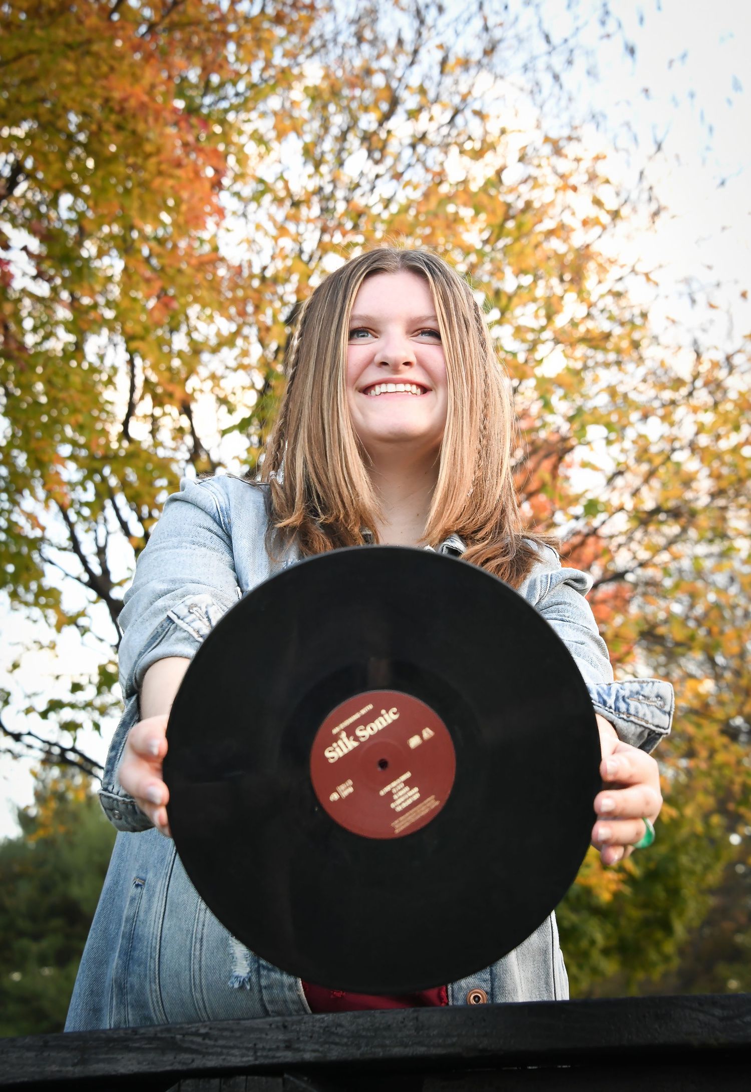Smiling girl holding vinyl record outdoors with colorful autumn trees.