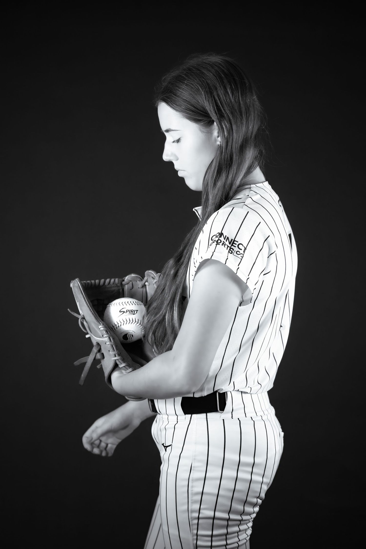Black and white photo of softball player in pinstripe uniform holding glove and ball in studio.