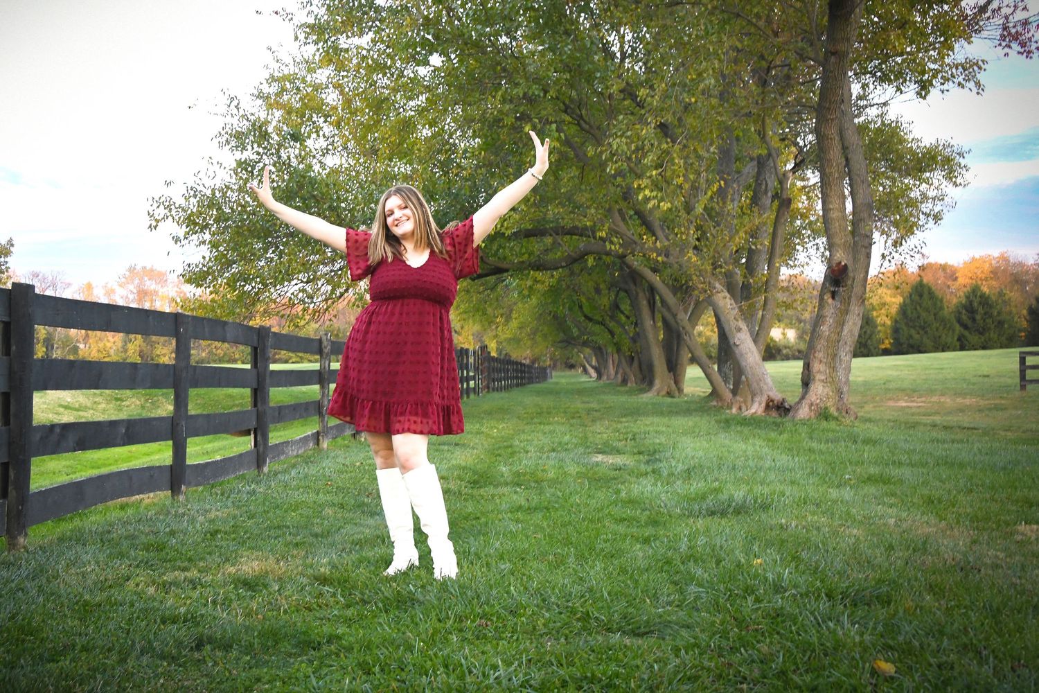 Joyful brunette girl in red dress and white boots raising arms outdoors by a wooden fence.