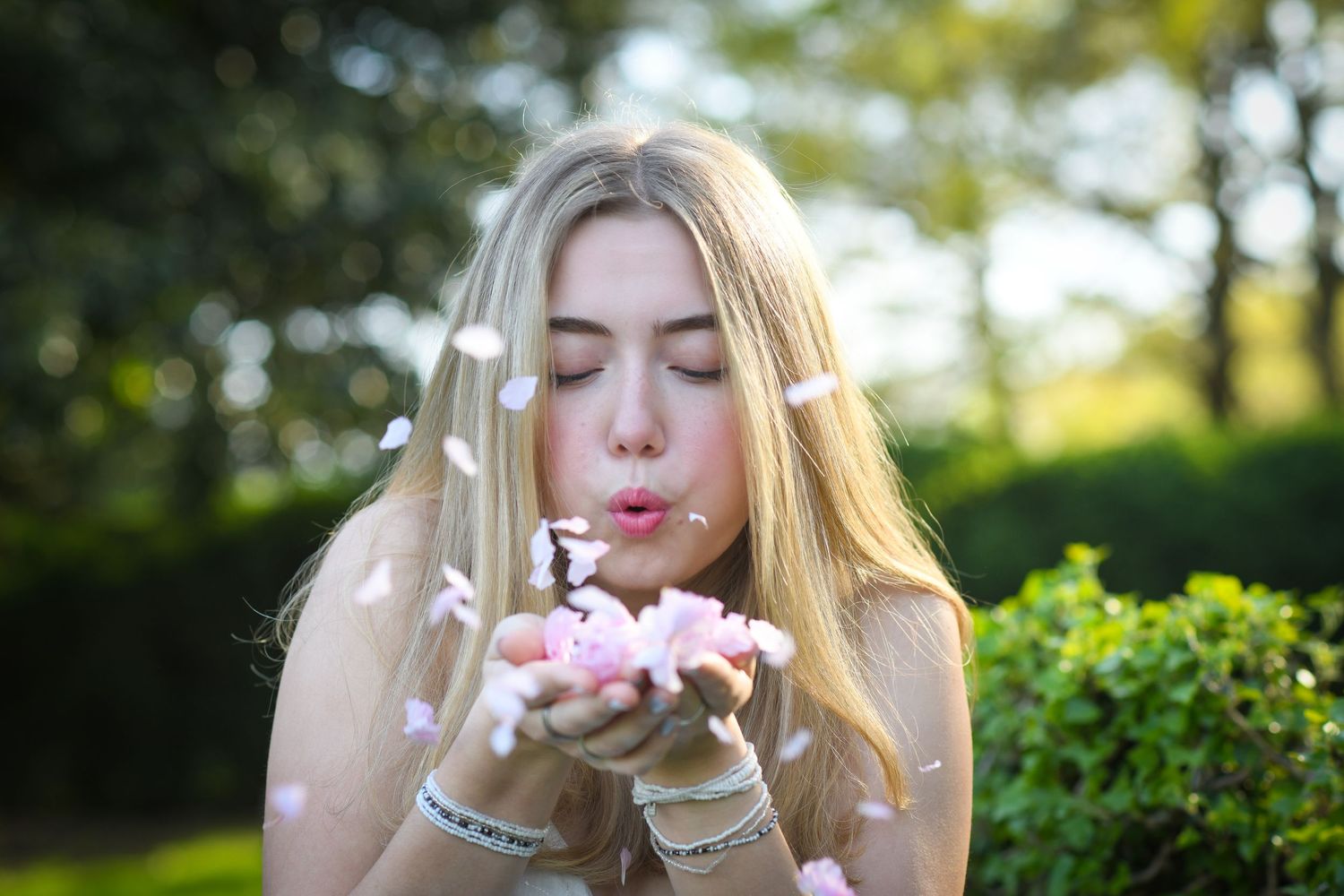 Young blonde woman blowing pink flower petals from her hands outdoors in a lush green garden setting.