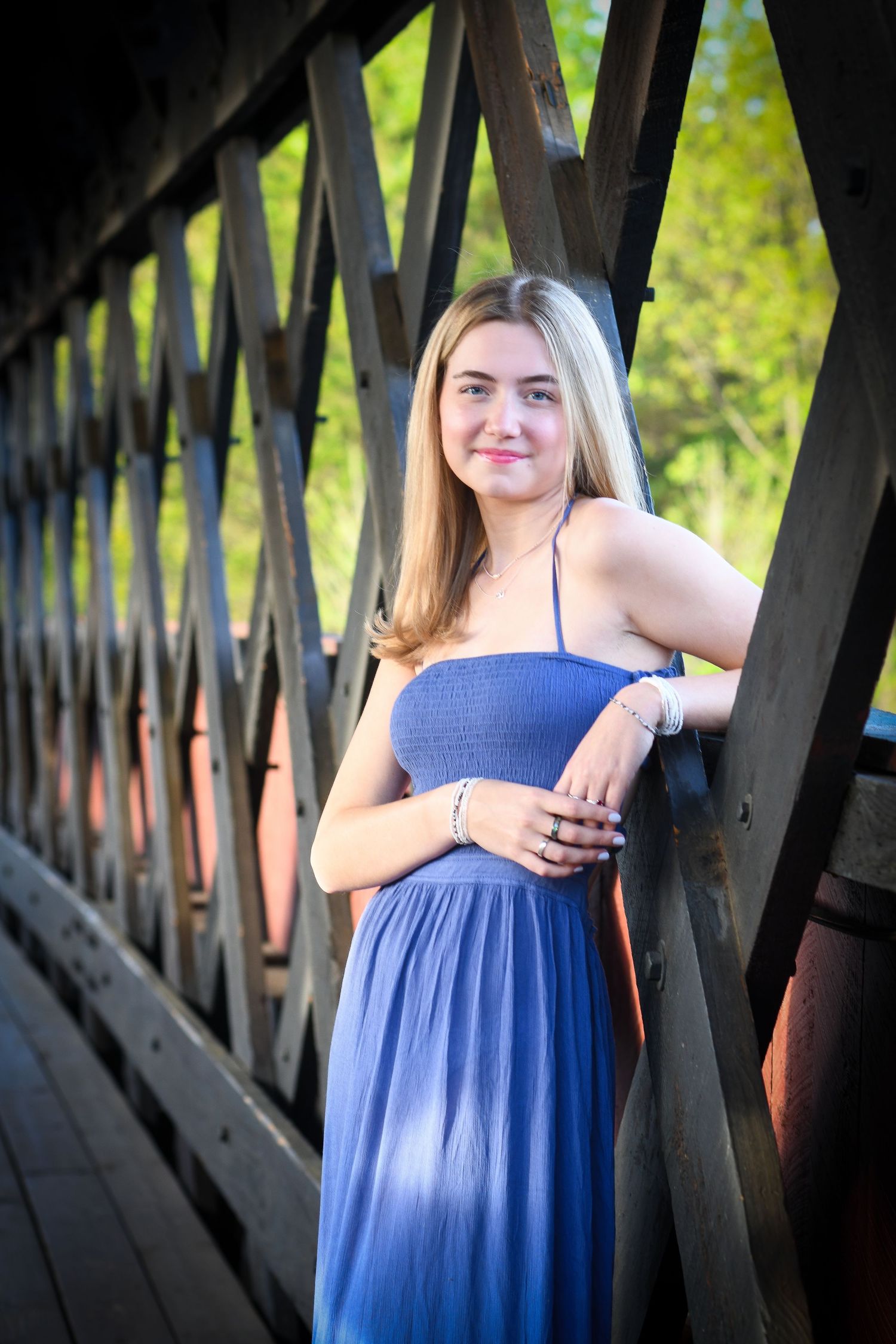 Smiling blonde teen in blue dress leaning against rustic wooden bridge structure outdoors.