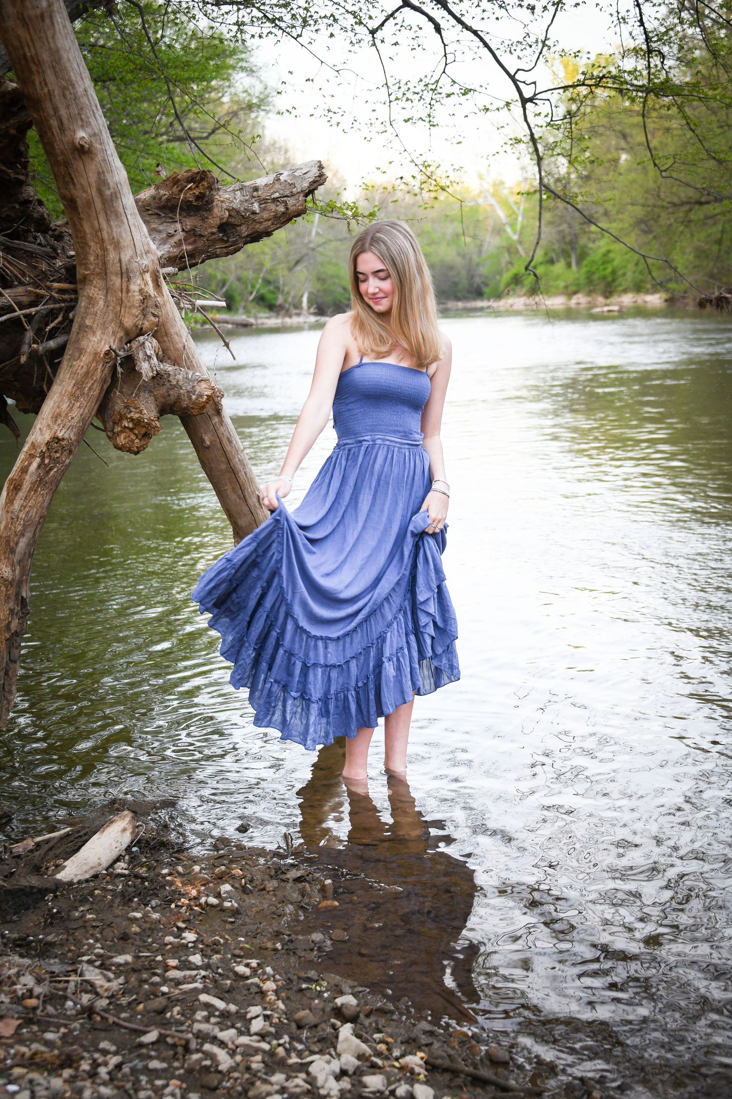 Blonde girl in blue strapless dress wading in river while holding skirt near fallen tree.
