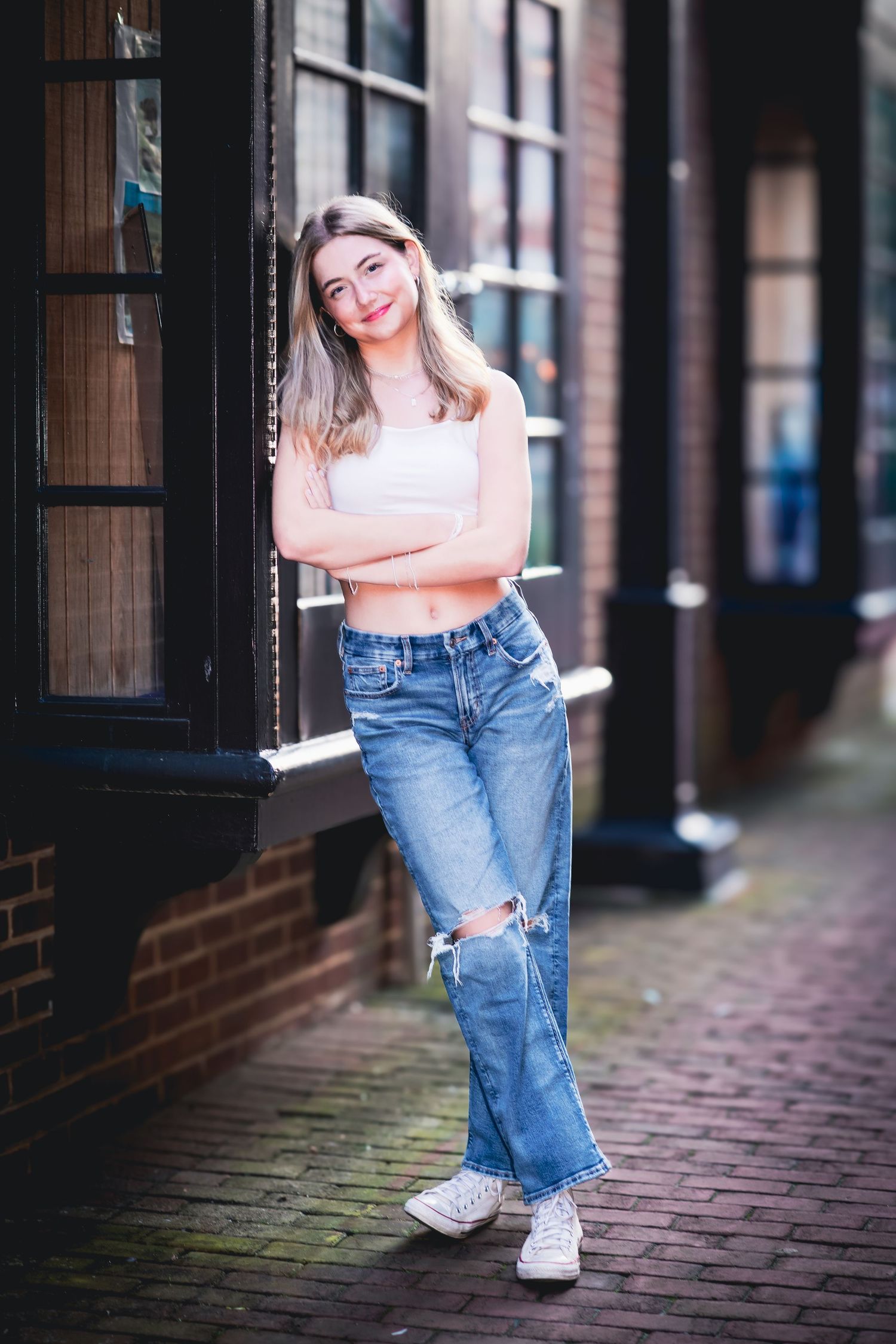 Smiling blonde teen in white crop top and ripped jeans posing on urban brick sidewalk.