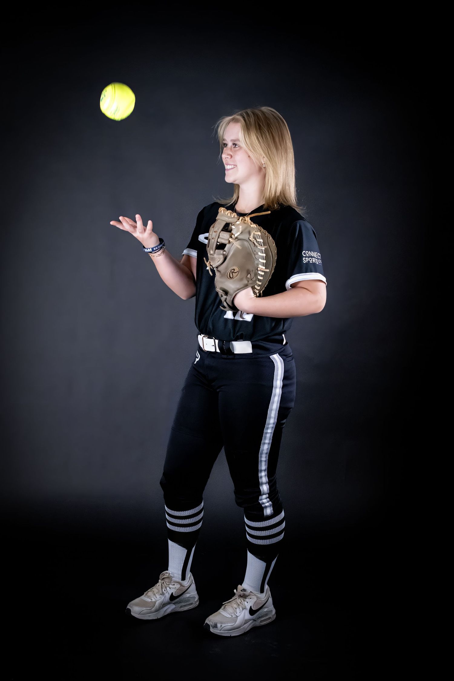 Blonde softball player in black uniform tossing yellow softball in studio.