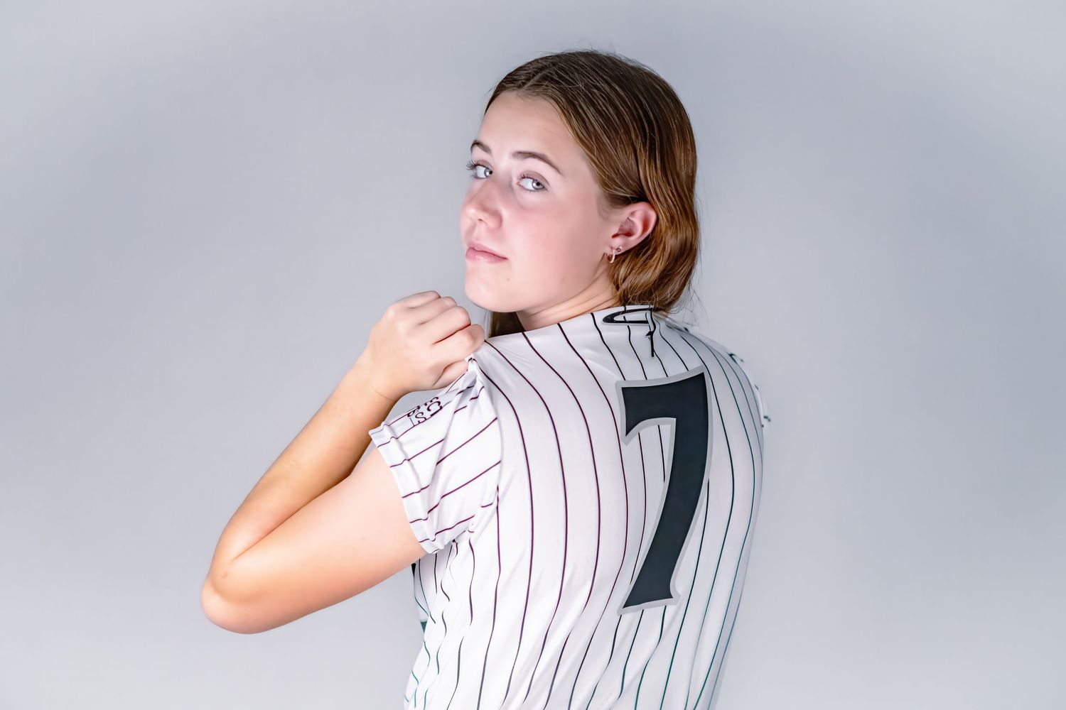 Young softball player in white pinstripe number 7 jersey looking over her shoulder in studio.