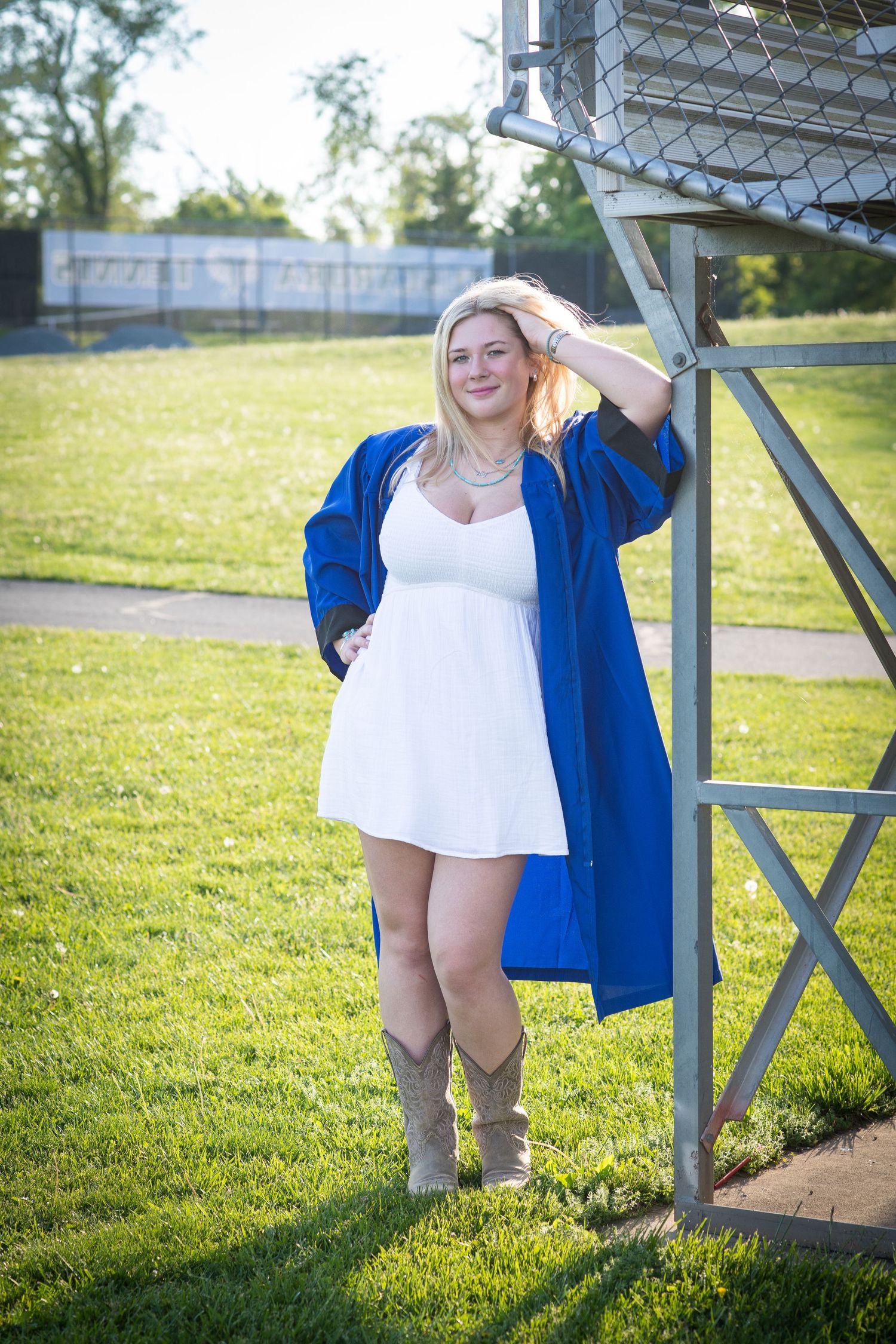 Smiling blonde graduate in blue cap and gown over white dress posing outdoors on grass.