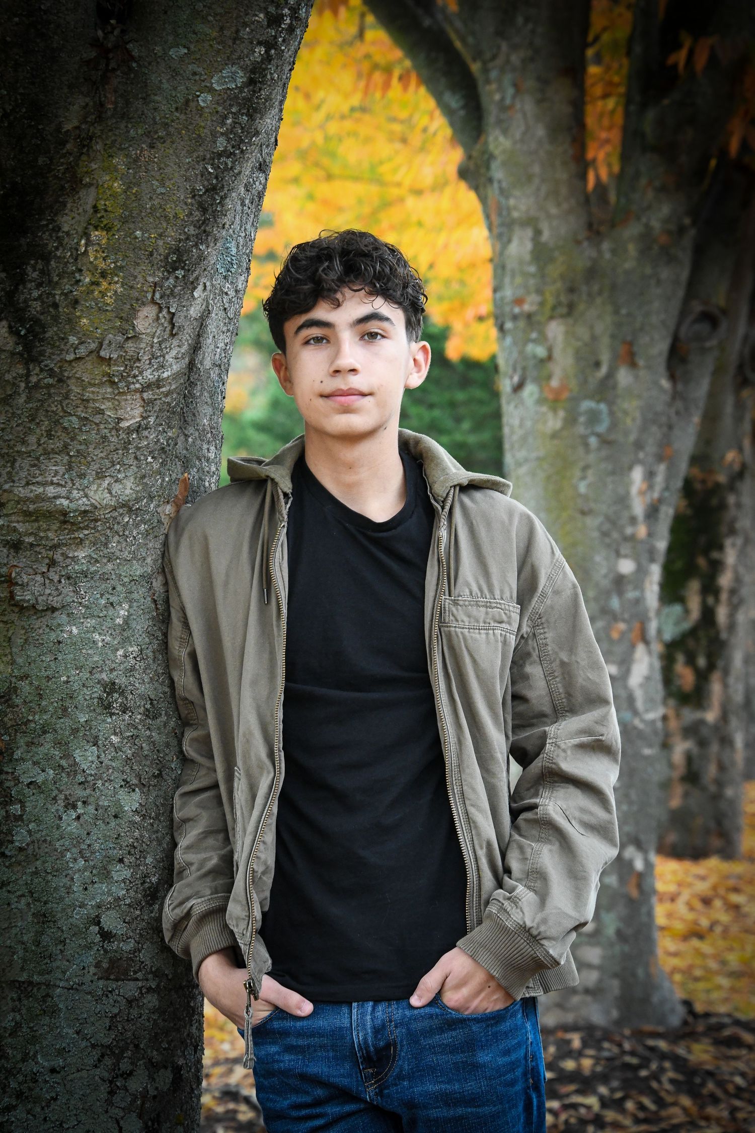 Teen boy in olive jacket and jeans leans against large tree with yellow autumn foliage backdrop.
