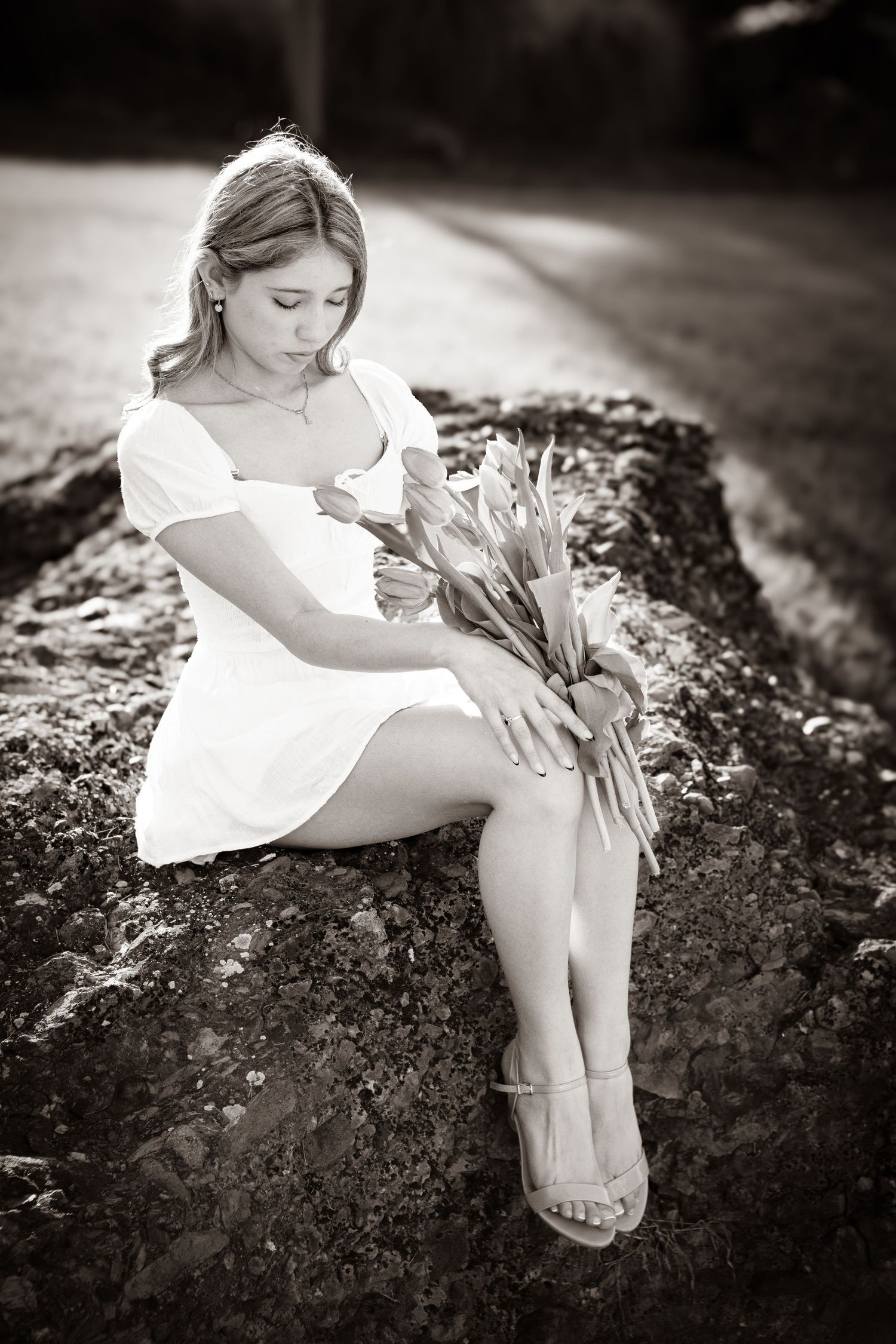 Young woman in white dress holding tulips while seated on stone ledge, elegant black and white portrait.