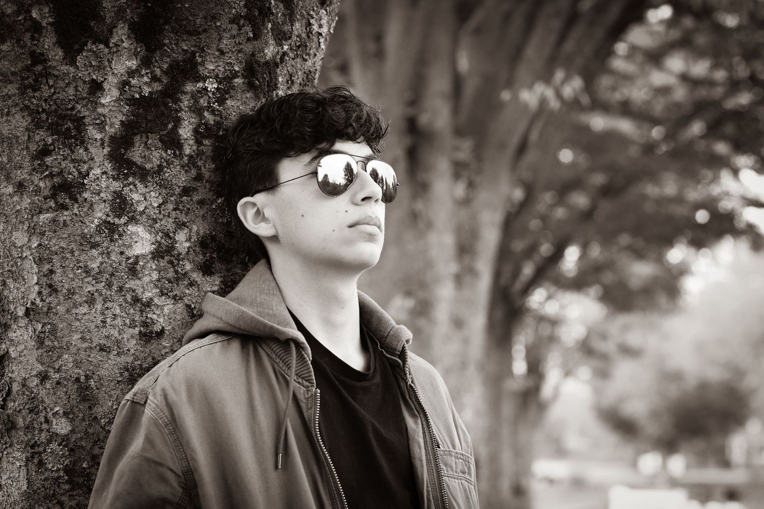 Black and white portrait of a young man with curly hair and aviator sunglasses leaning on a tree.