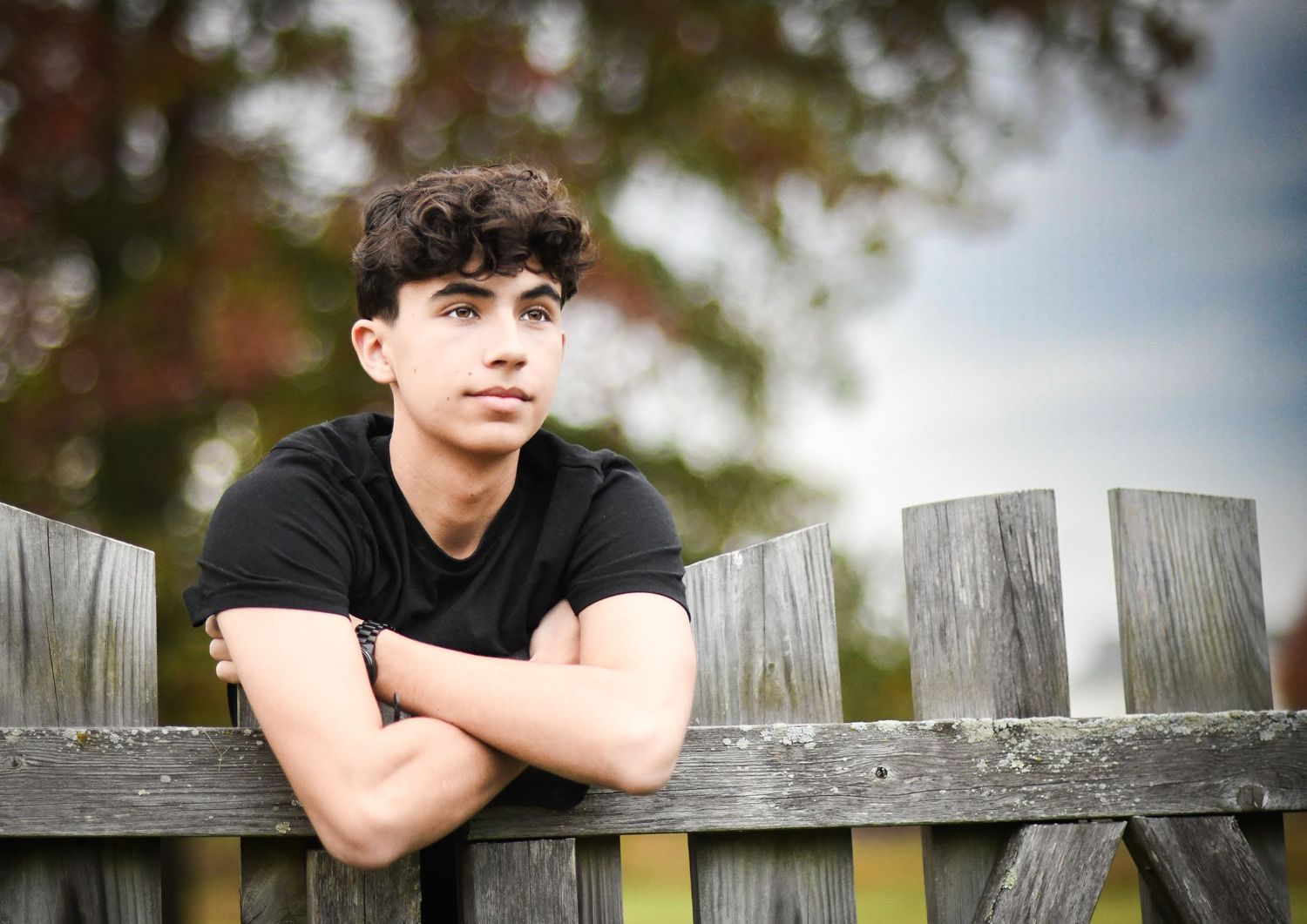Teen boy with curly hair leaning on wooden fence with arms crossed in outdoor portrait.