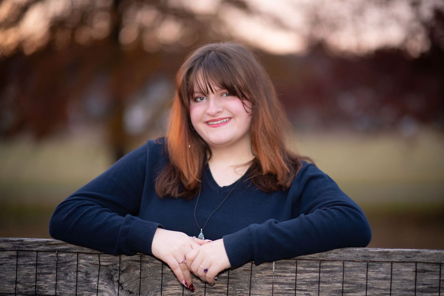 Smiling young woman with bangs leaning on a wooden fence during outdoor portrait session.