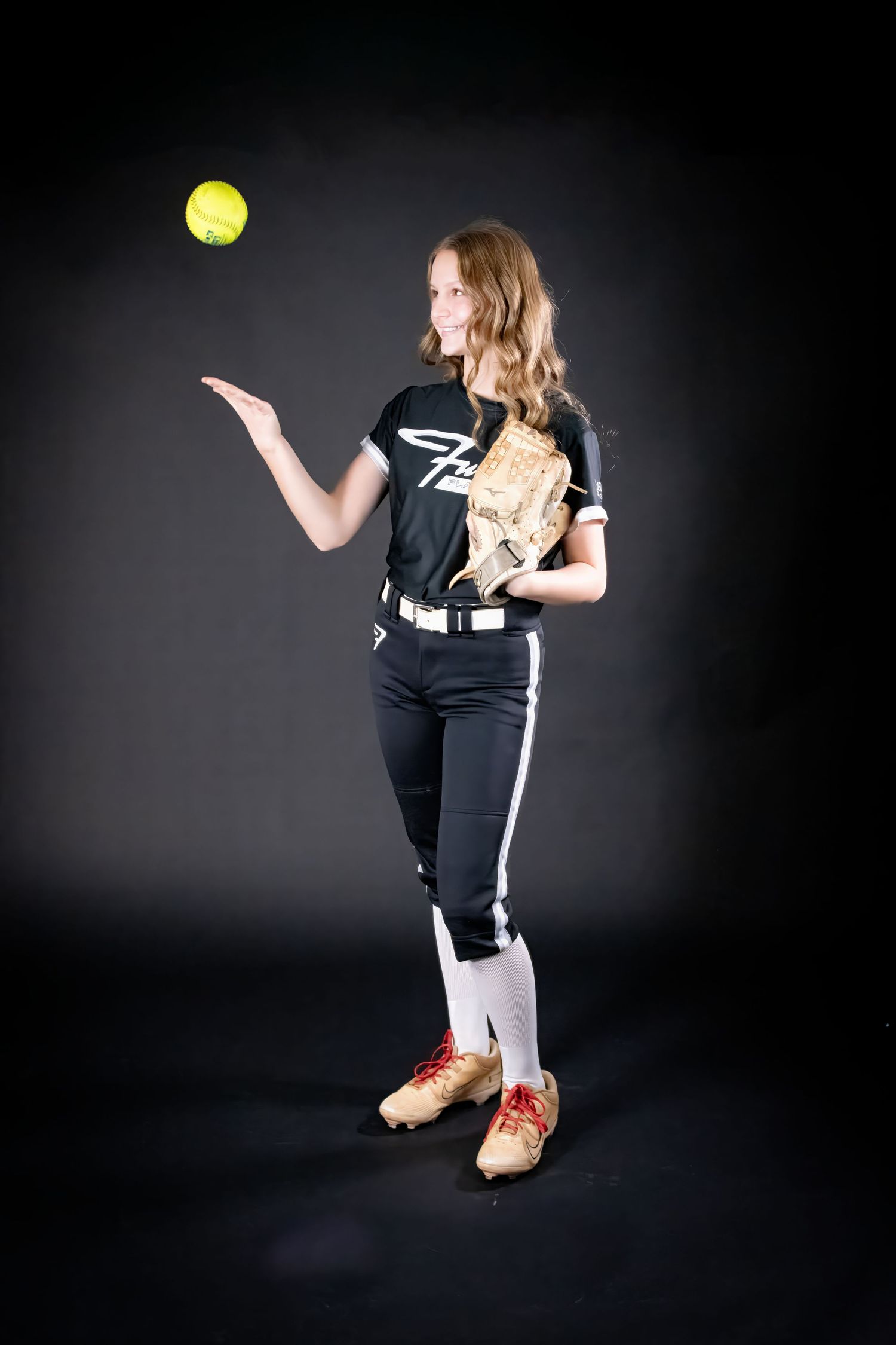 Female softball player in black uniform tossing yellow ball with glove in hand.