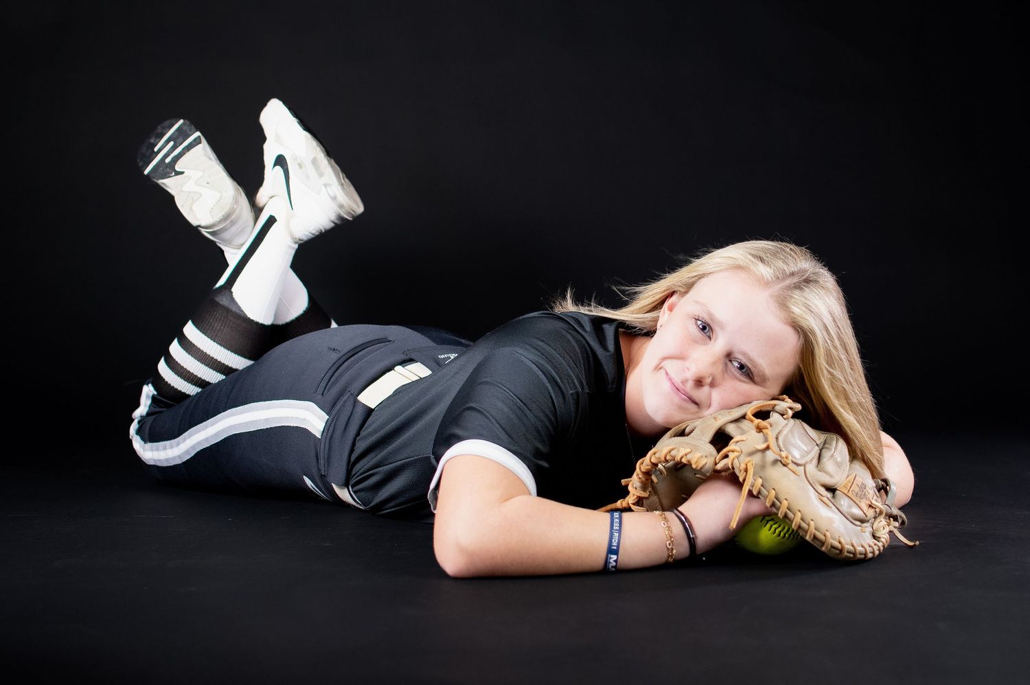 Smiling blonde female softball player lying down with glove and ball in black uniform.