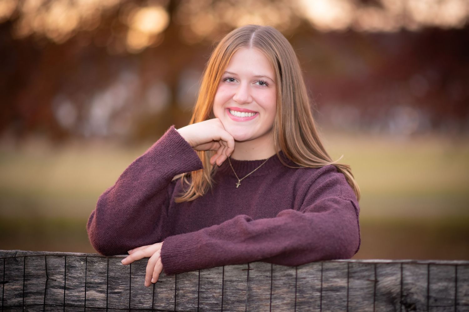 Young woman with long hair smiling while leaning on a fence at golden hour.