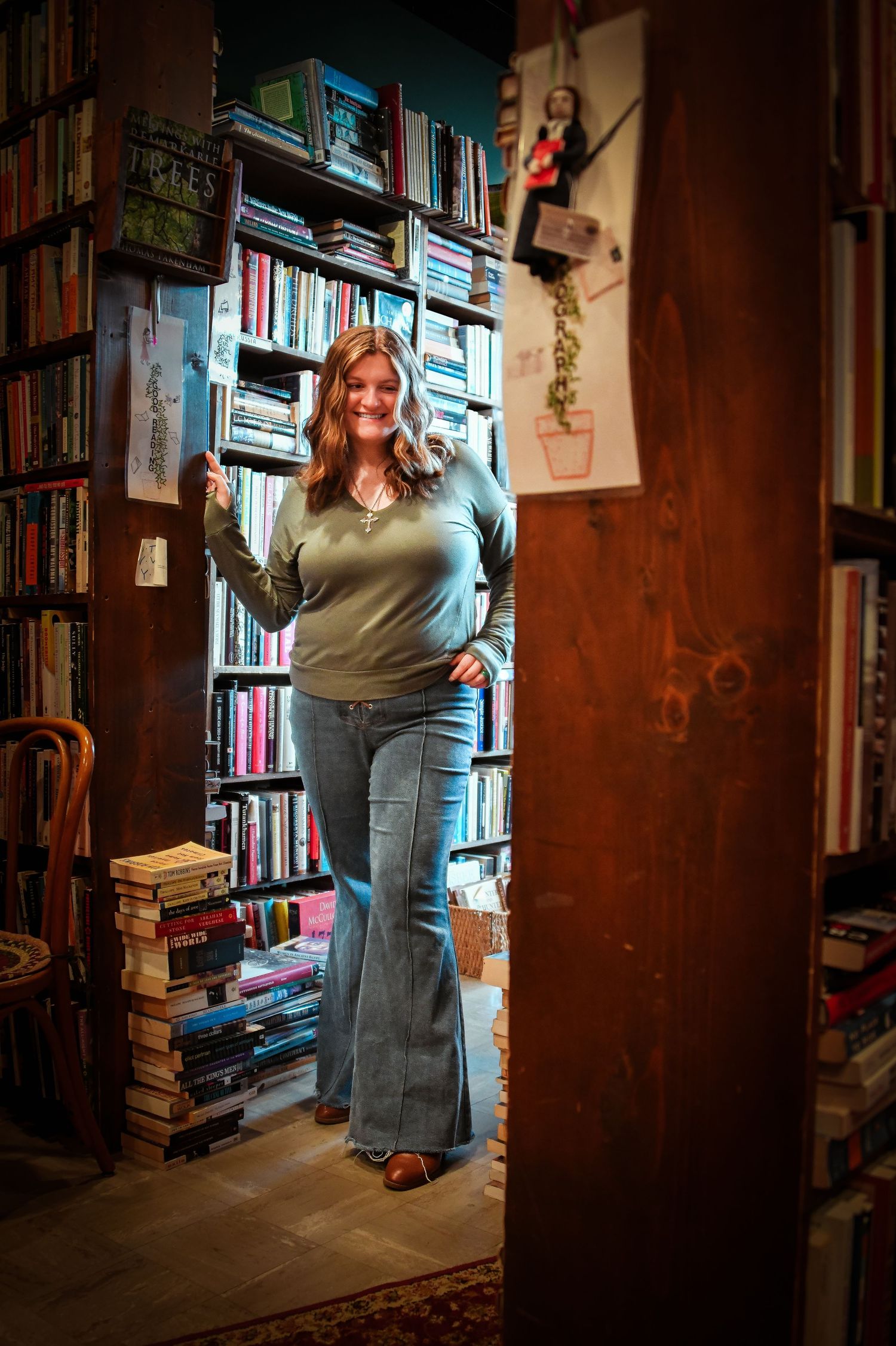 Curvy woman in green sweater and flare jeans poses in doorway of eclectic bookstore interior.