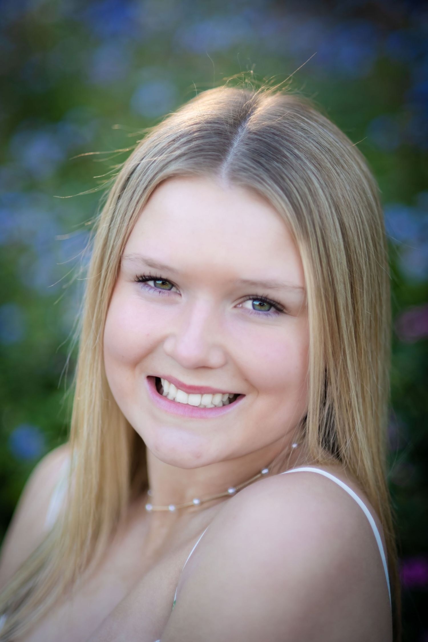 Smiling blonde teenage girl with blue eyes posing in front of purple flowers for a senior portrait.