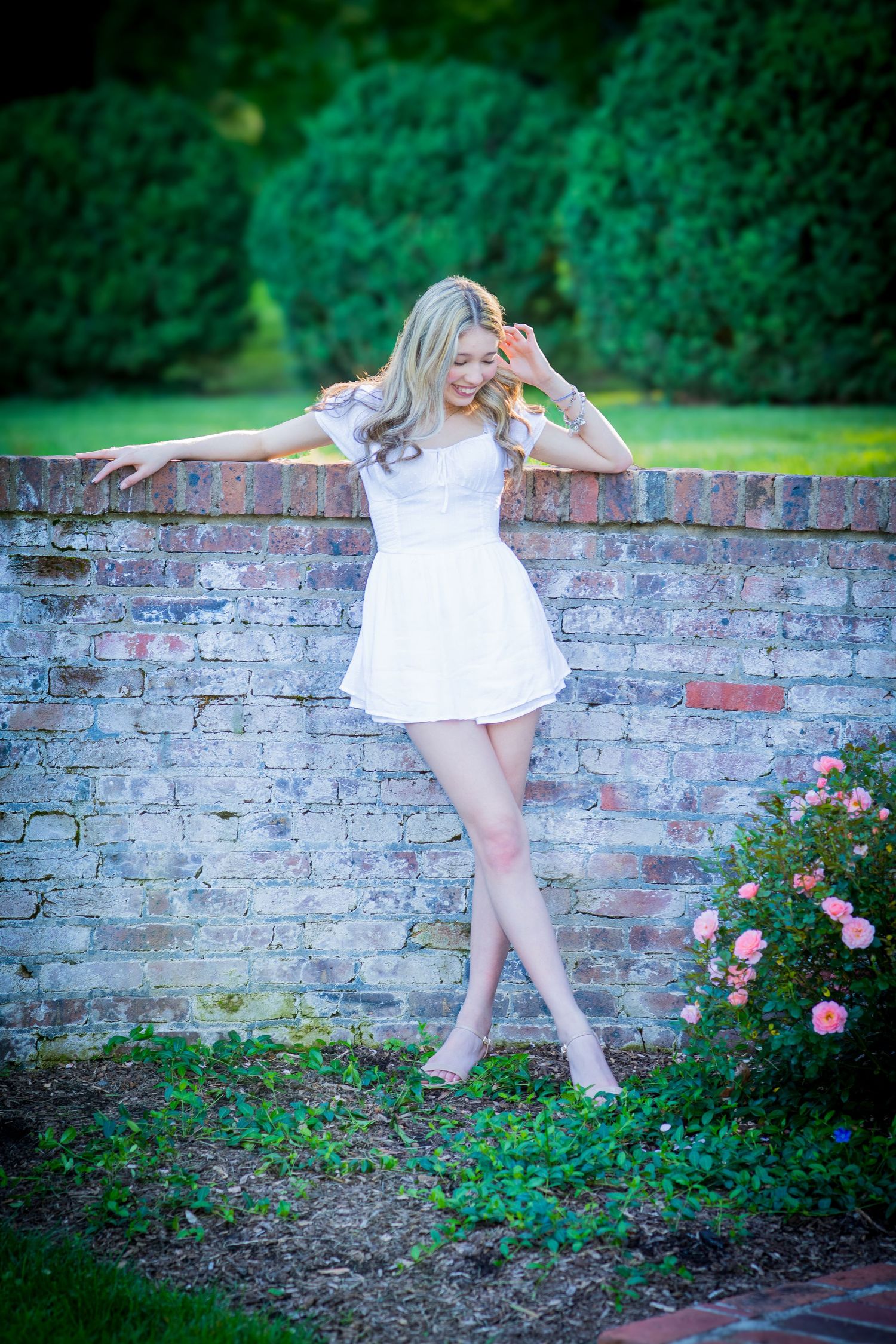 Blonde teen girl in white mini dress poses gracefully against rustic brick wall with roses nearby.