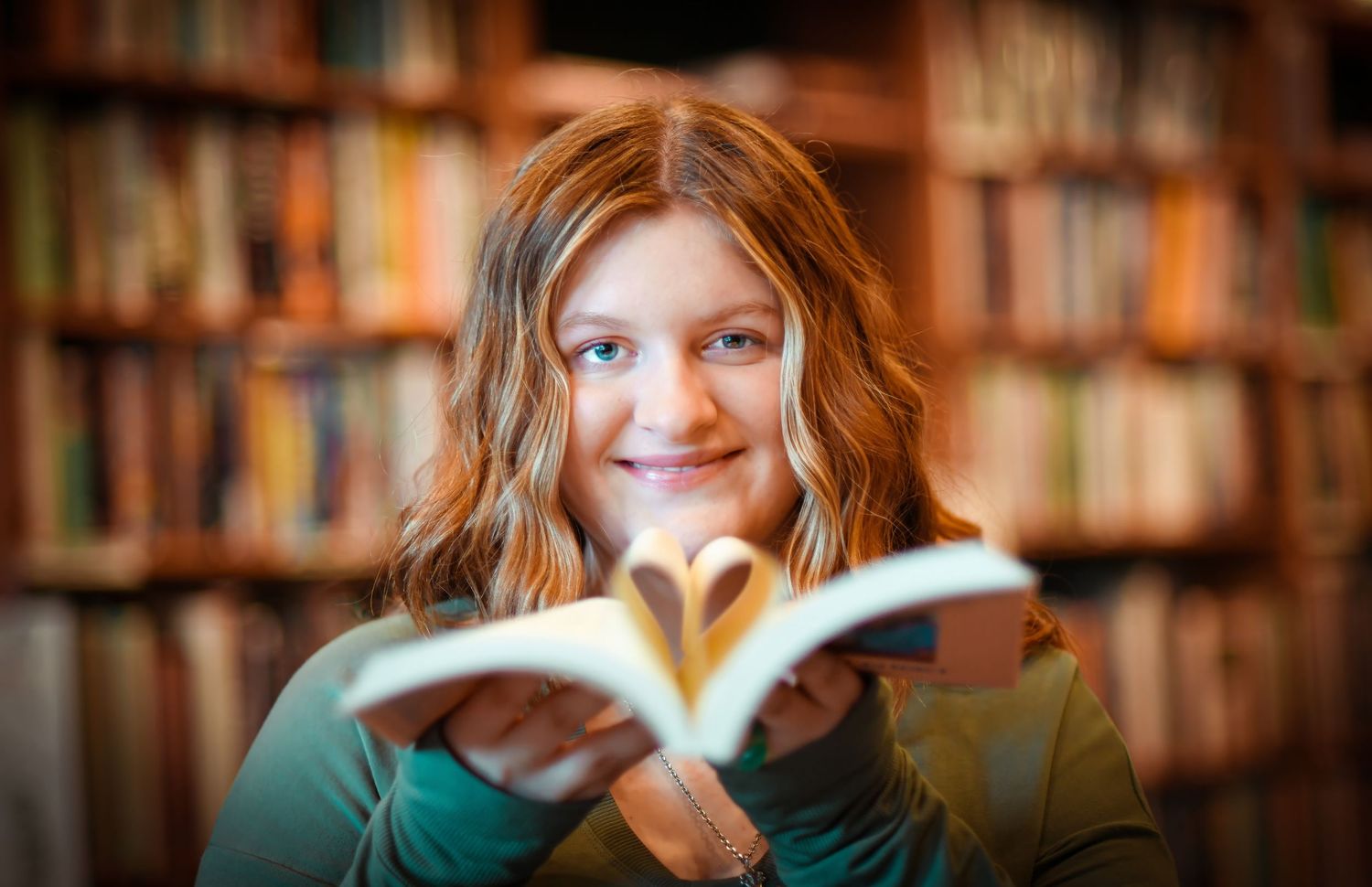 Smiling young girl holds open book forming heart shape in front of colorful library bookshelves.