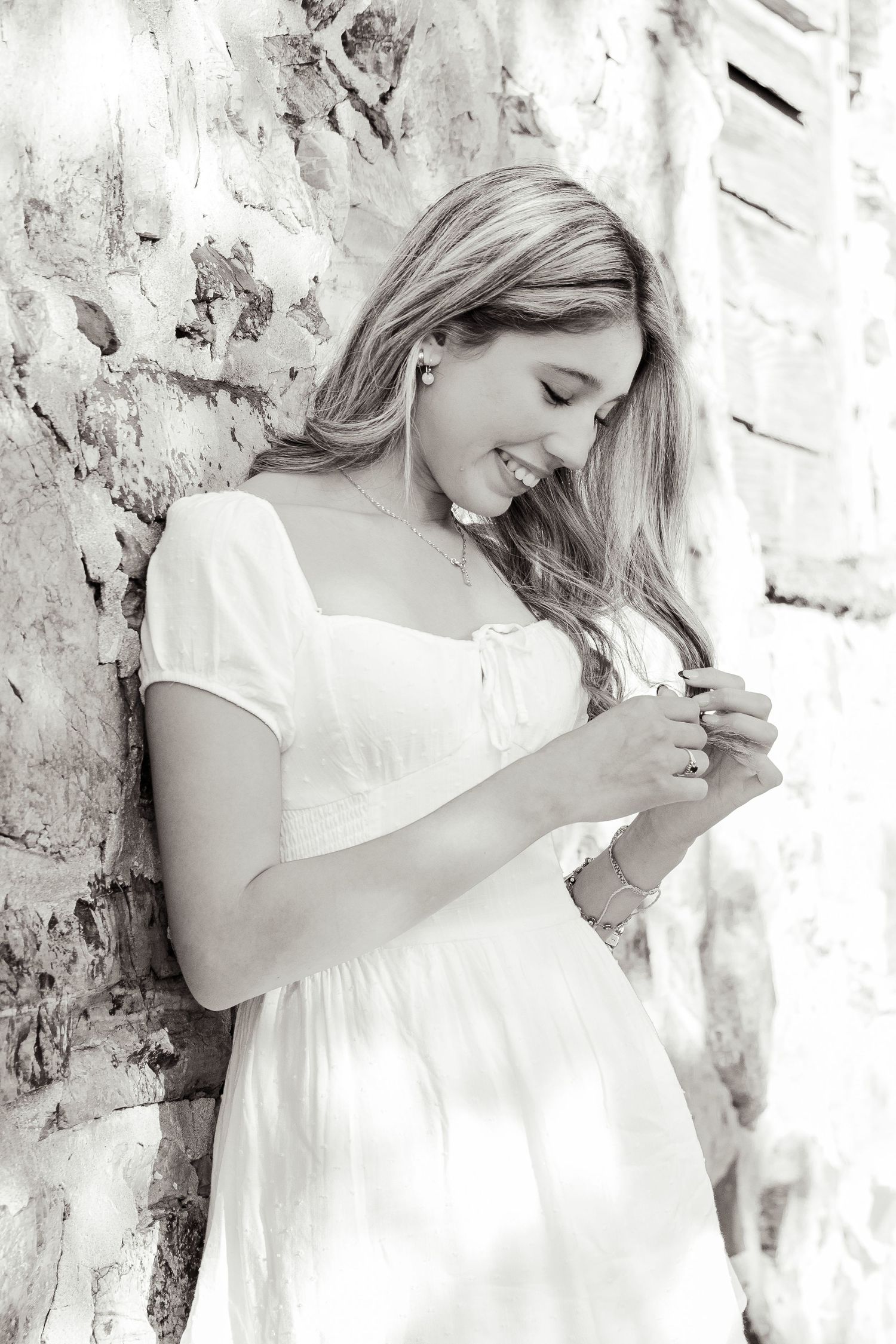 Blonde woman in white dress leaning against stone wall smiling downward, black and white senior portrait.