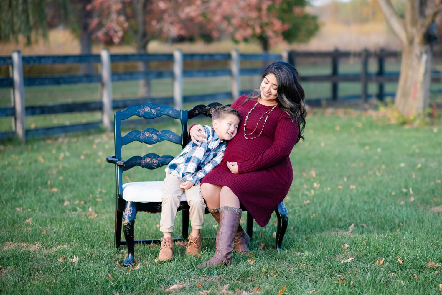 Pregnant woman in burgundy dress sitting on bench with young boy during fall maternity session.