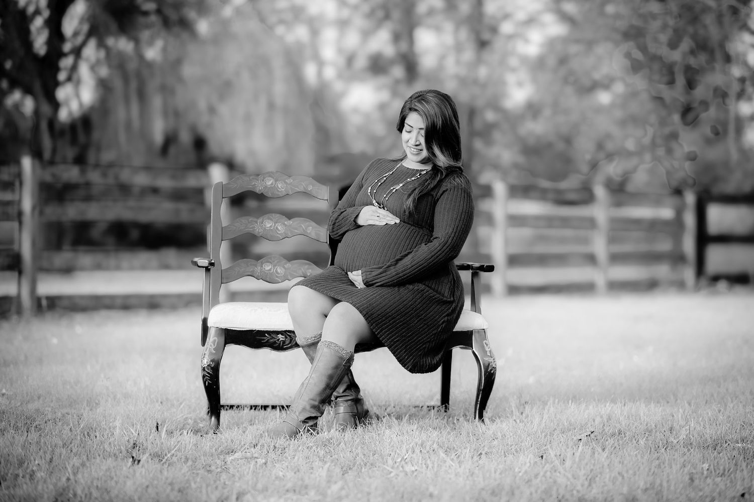 Black and white photo of pregnant woman sitting on bench in field wearing boots and dress.