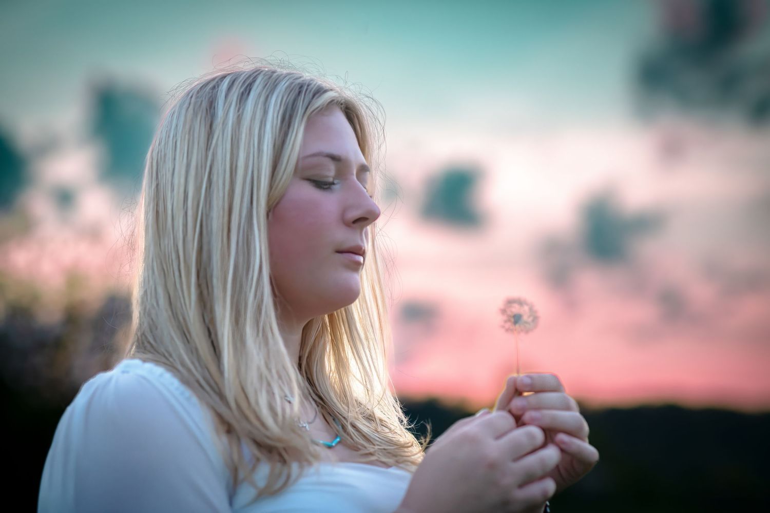 Young blonde woman blowing dandelion seeds at sunset in white top.