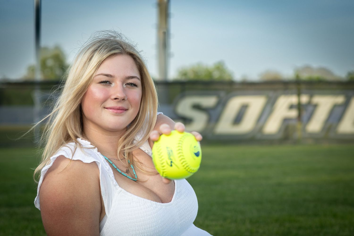 Blonde softball player in white top holding yellow softball on field with Softball sign.